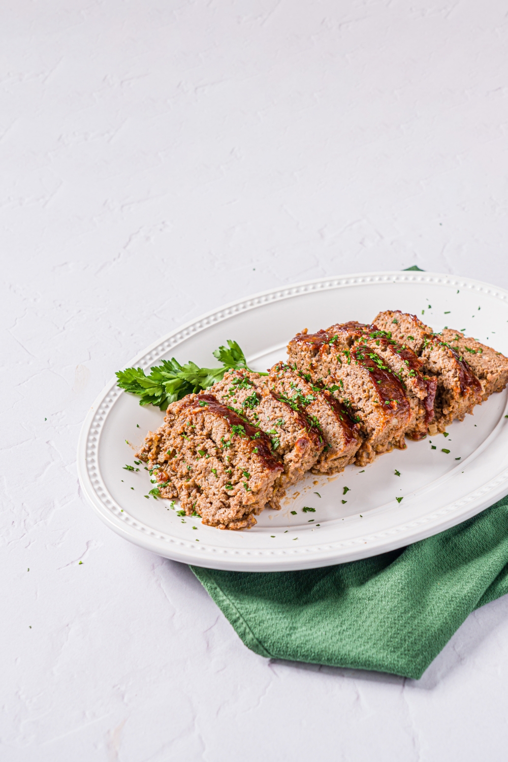 A white platter with sliced meatloaf topped with glaze and garnished with fresh parsley. The platter is on a marble counter with a green cloth napkin.