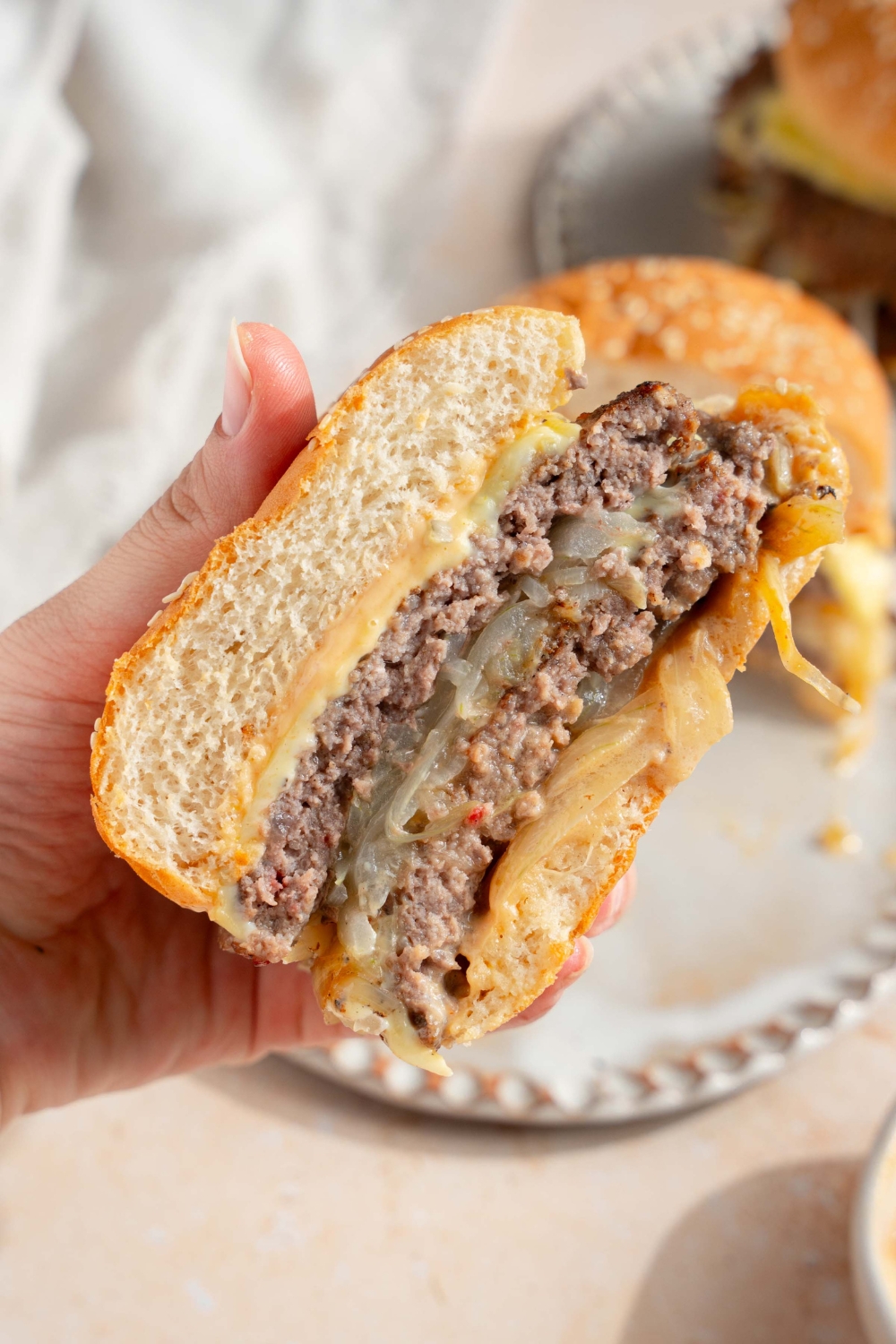 A close up of a hand holding a smash burger with onions sliced in half. There is a plate with the remaining burger on a tan counter blurred in the background.