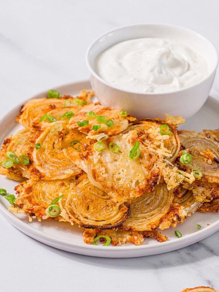 A white plate with baked parmesan onion chips garnished with sliced green onions and small bowl of dip. The plate is on a marble counter.