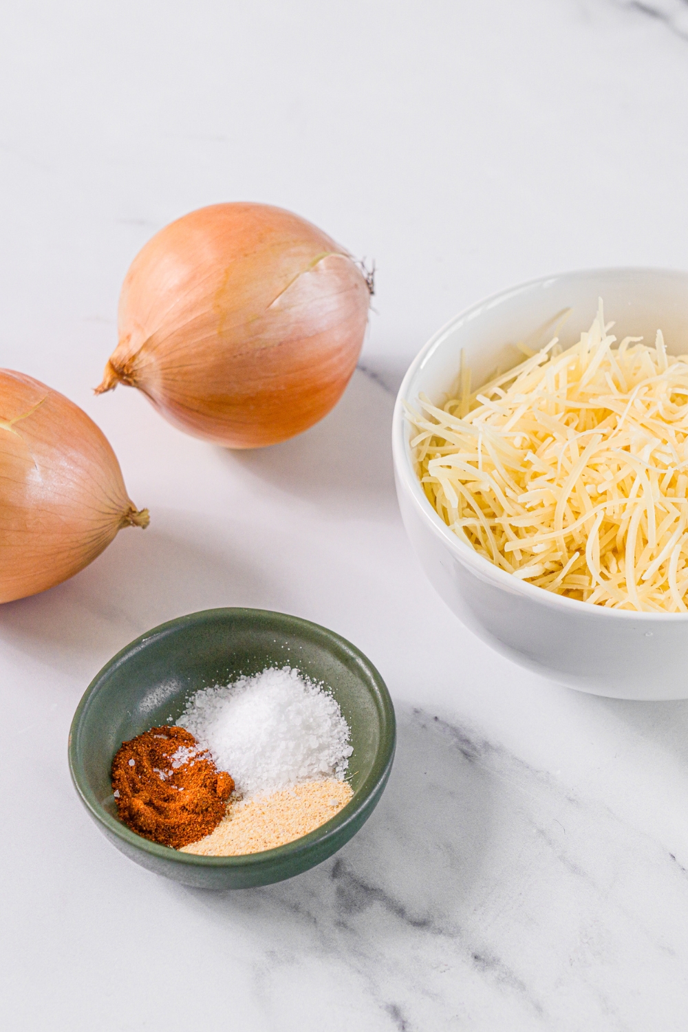 A marble counter with ingredients to make onion chips including white onion, shredded parmesan cheese, and a bowl of seasonings.