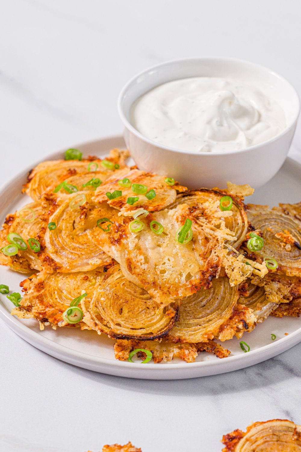 A white plate with baked parmesan onion chips garnished with sliced green onions and small bowl of dip. The plate is on a marble counter.