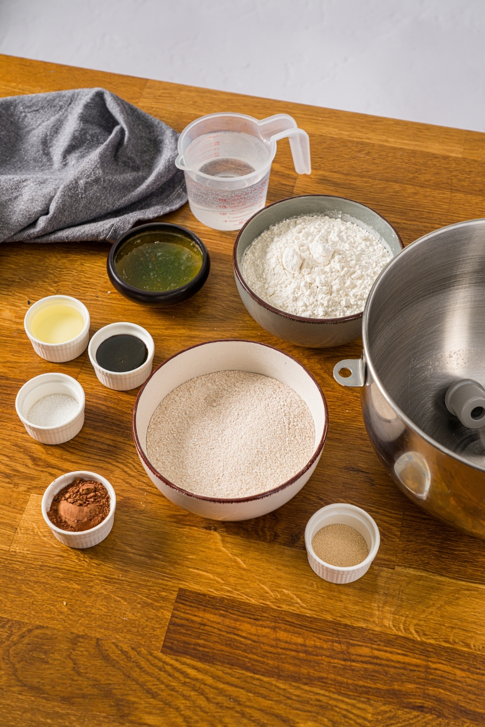 A wooden board with several bowls in various sizes containing ingredients to make Outback sweet molasses brown bread including flour, whole wheat flour, molasses, cocoa powder, yeast, oil, water, and honey.