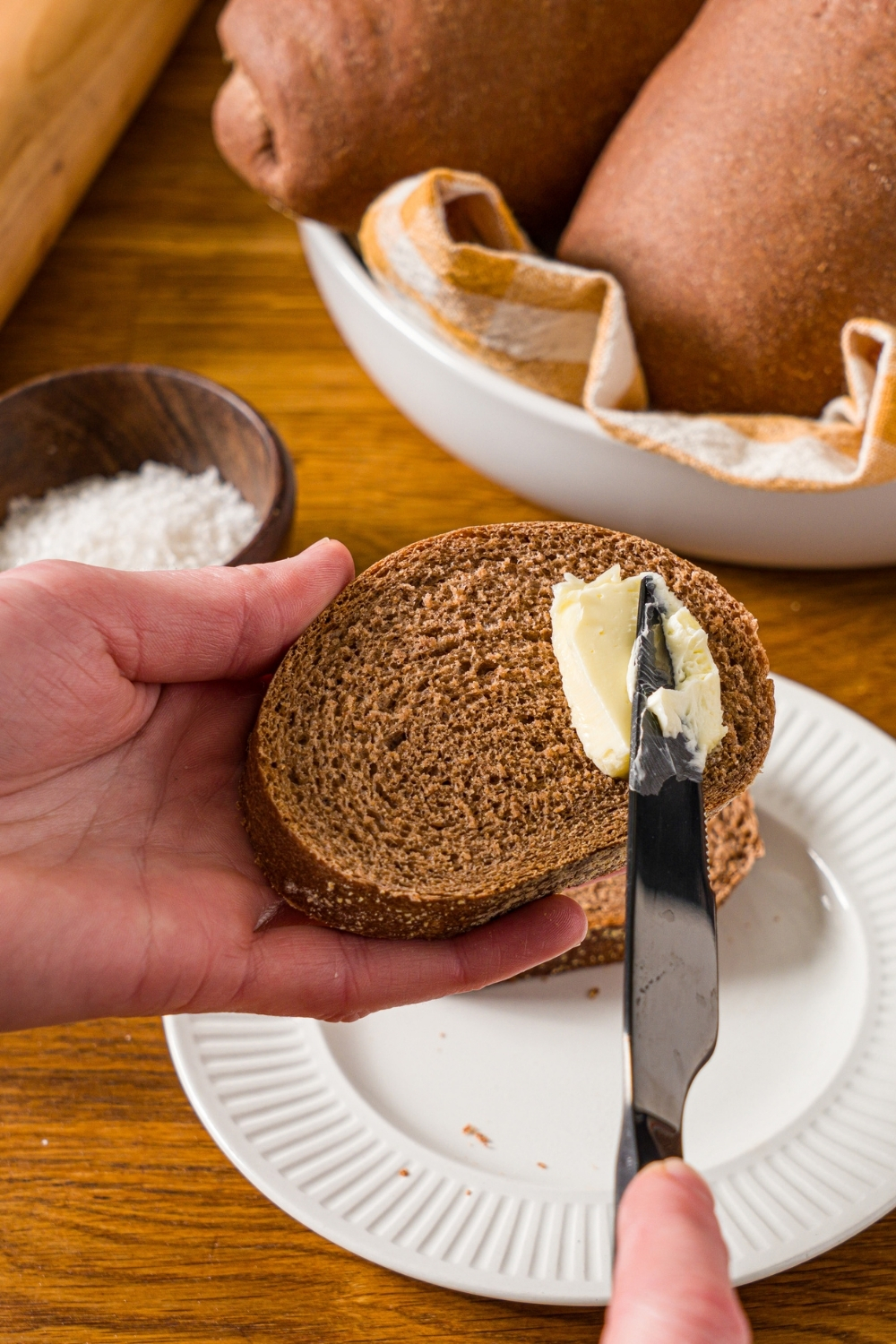 A hand buttering a slice of Outback sweet molasses brown bread with a knife. There is a white plate with another slice of bread in the background. The plate is on a wooden counter with a bowl of bread loaves.