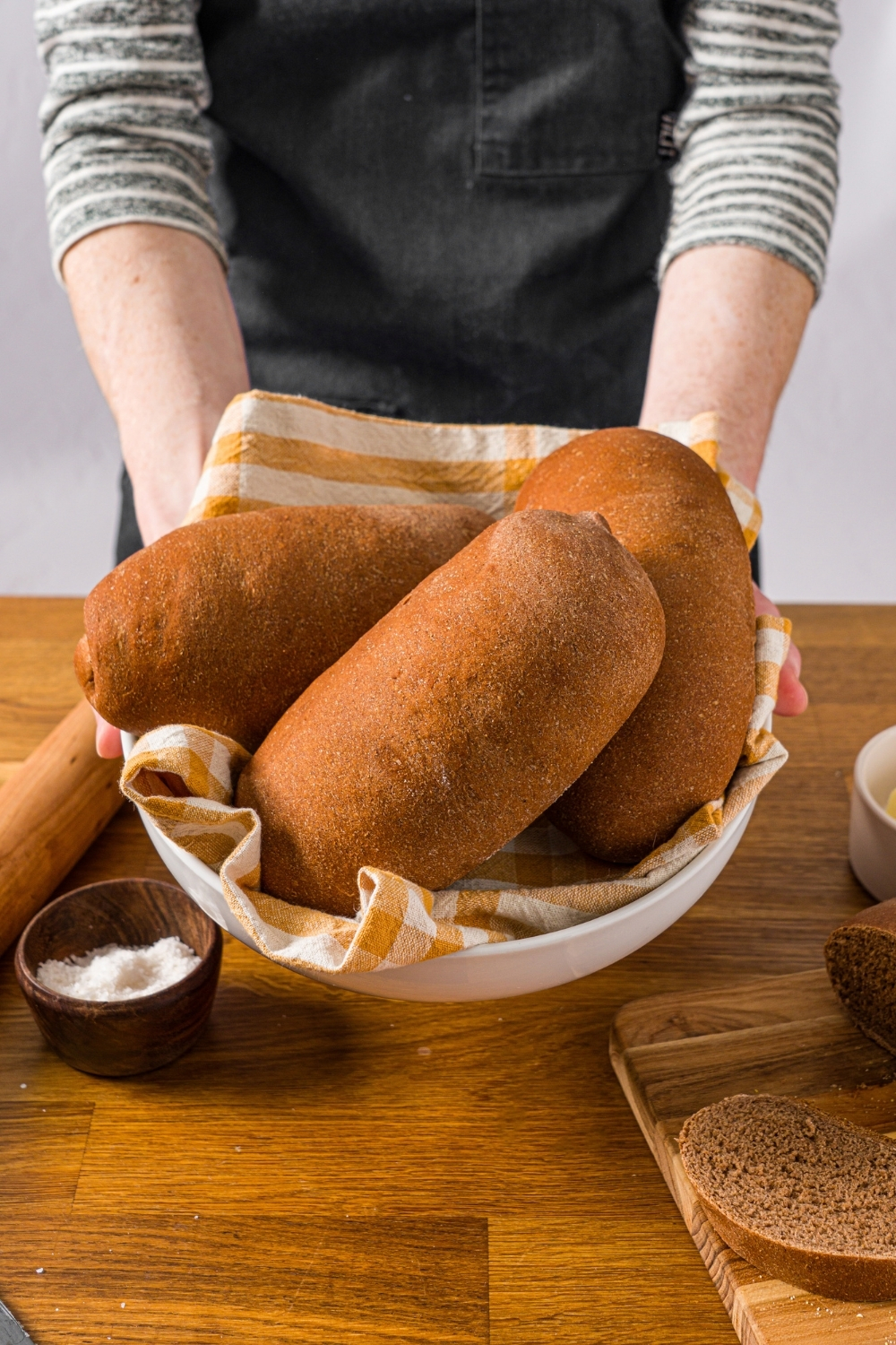 Hands holding a large bowl of Outback sweet molasses brown bread loaves. The bowl is lined with a yellow striped napkin. The bowl is over a wooden counter.