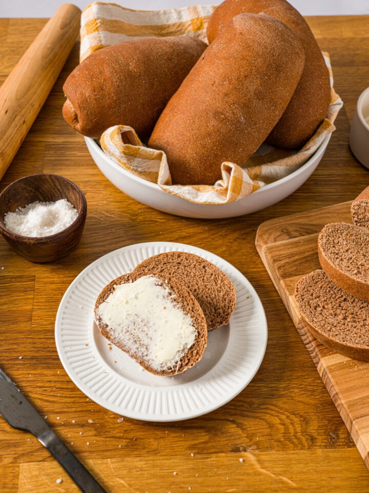 A white plate with two slices of Outback sweet molasses brown bread. One of the slices is buttered. The plate is on a wooden counter with a bowl of loaves and a cutting board with additional slices of bread.