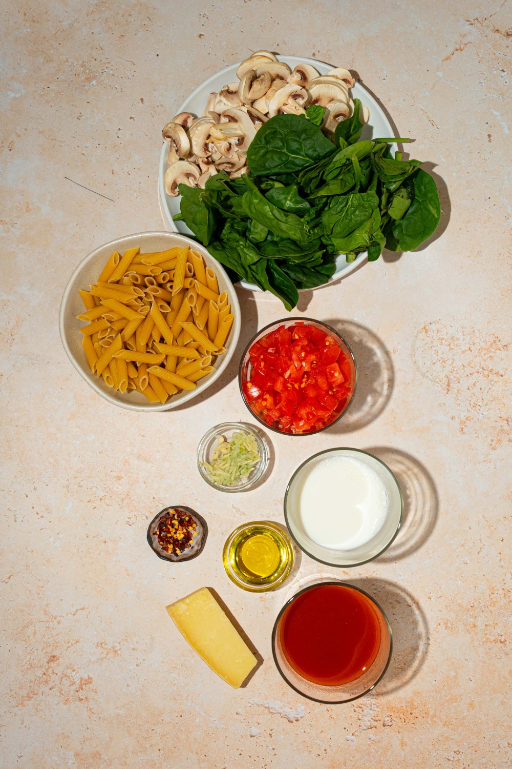 An overhead shot of several bowls in various sizes containing ingredients to make penne rosa including penne pasta, spinach, marinara sauce, mushrooms, garlic, crushed tomatoes, oil, cream, and red pepper flakes.