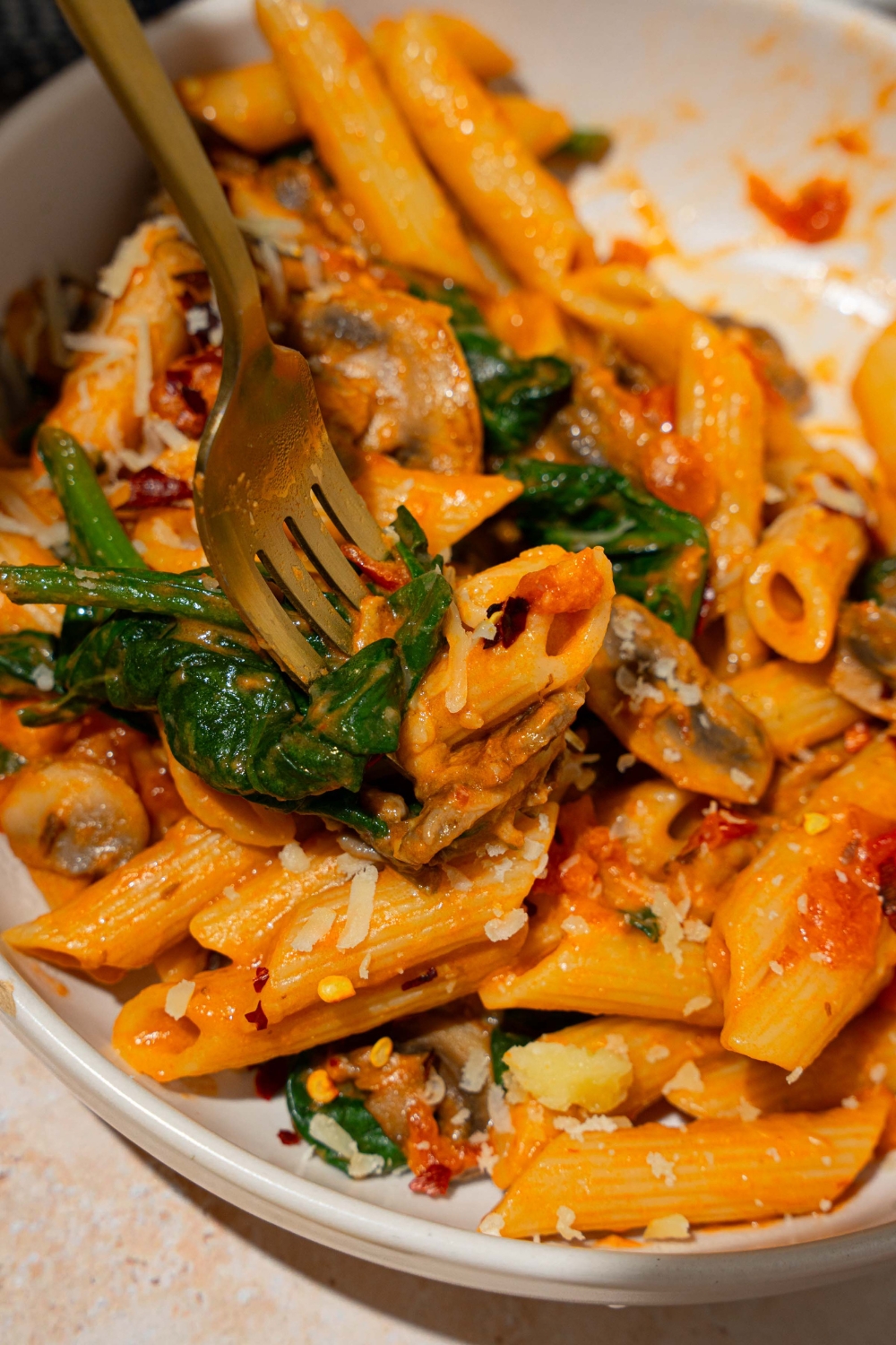 A white plate with penne rosa tossed in spinach and mushrooms. There is a fork taking a bite from the plate.