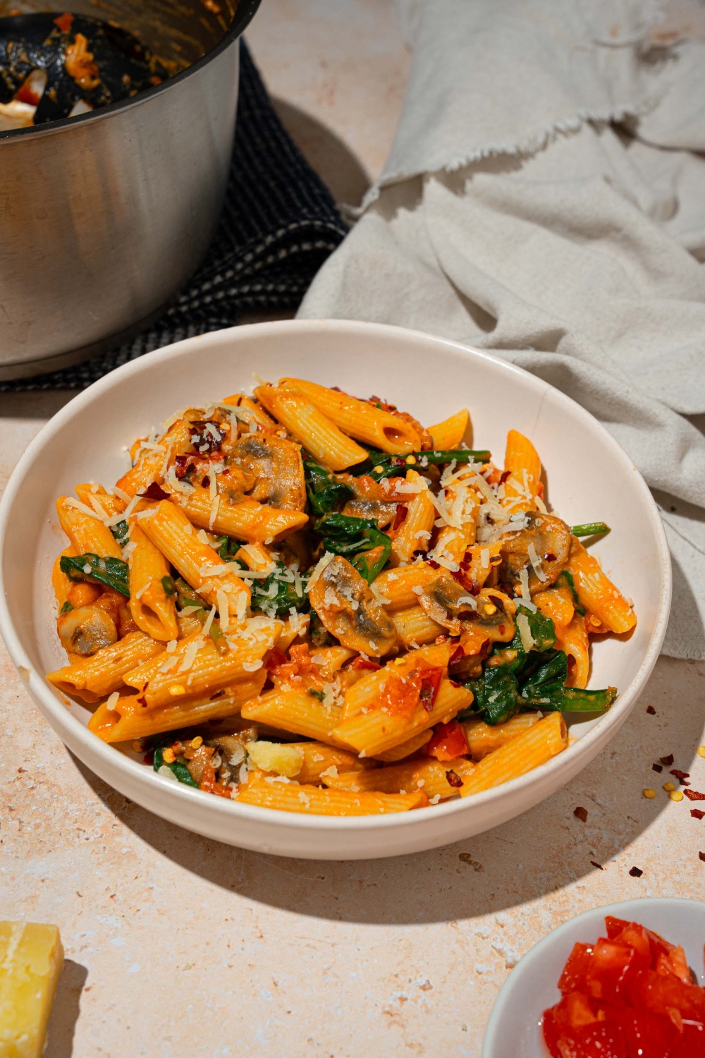 A white plate with penne rosa tossed in spinach and mushrooms and garnished with shredded parmesan. There is a fork taking a bite from the plate. The plate is on a tan counter with a white cloth napkin and pot of penne rosa.