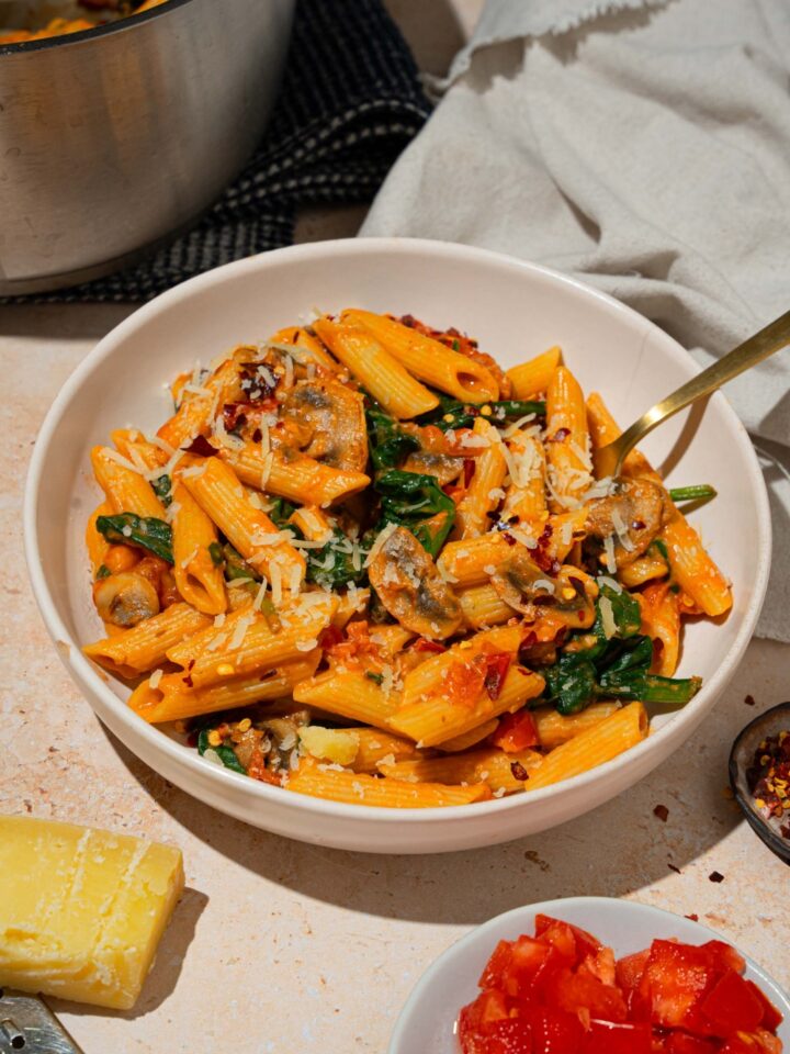 A white plate with penne rosa tossed in spinach and mushrooms and garnished with shredded parmesan. There is a fork in the bowl. The plate is on a tan counter with a white cloth napkin and pot of penne rosa.