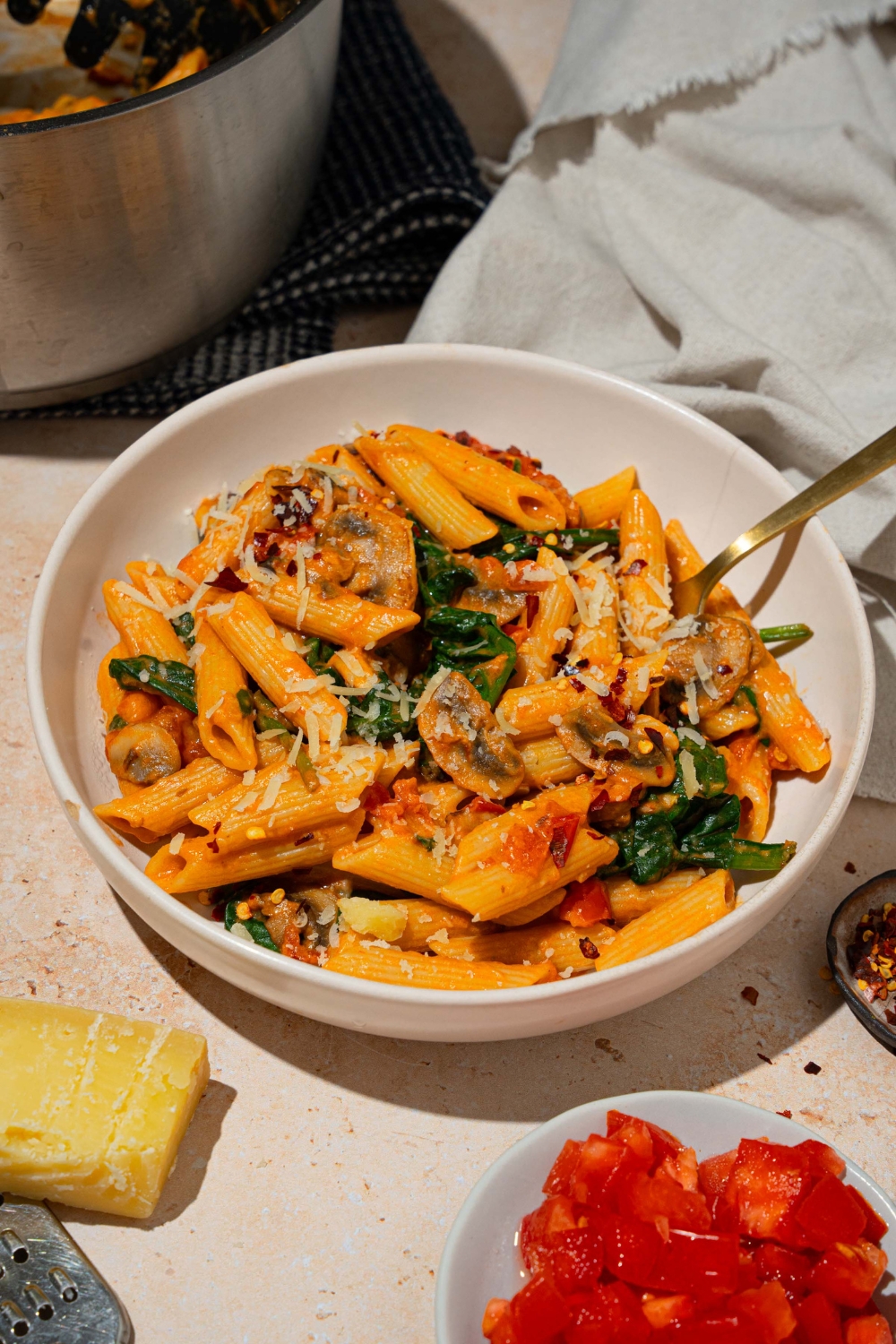 A white plate with penne rosa tossed in spinach and mushrooms and garnished with shredded parmesan. There is a fork in the bowl. The plate is on a tan counter with a white cloth napkin and pot of penne rosa.