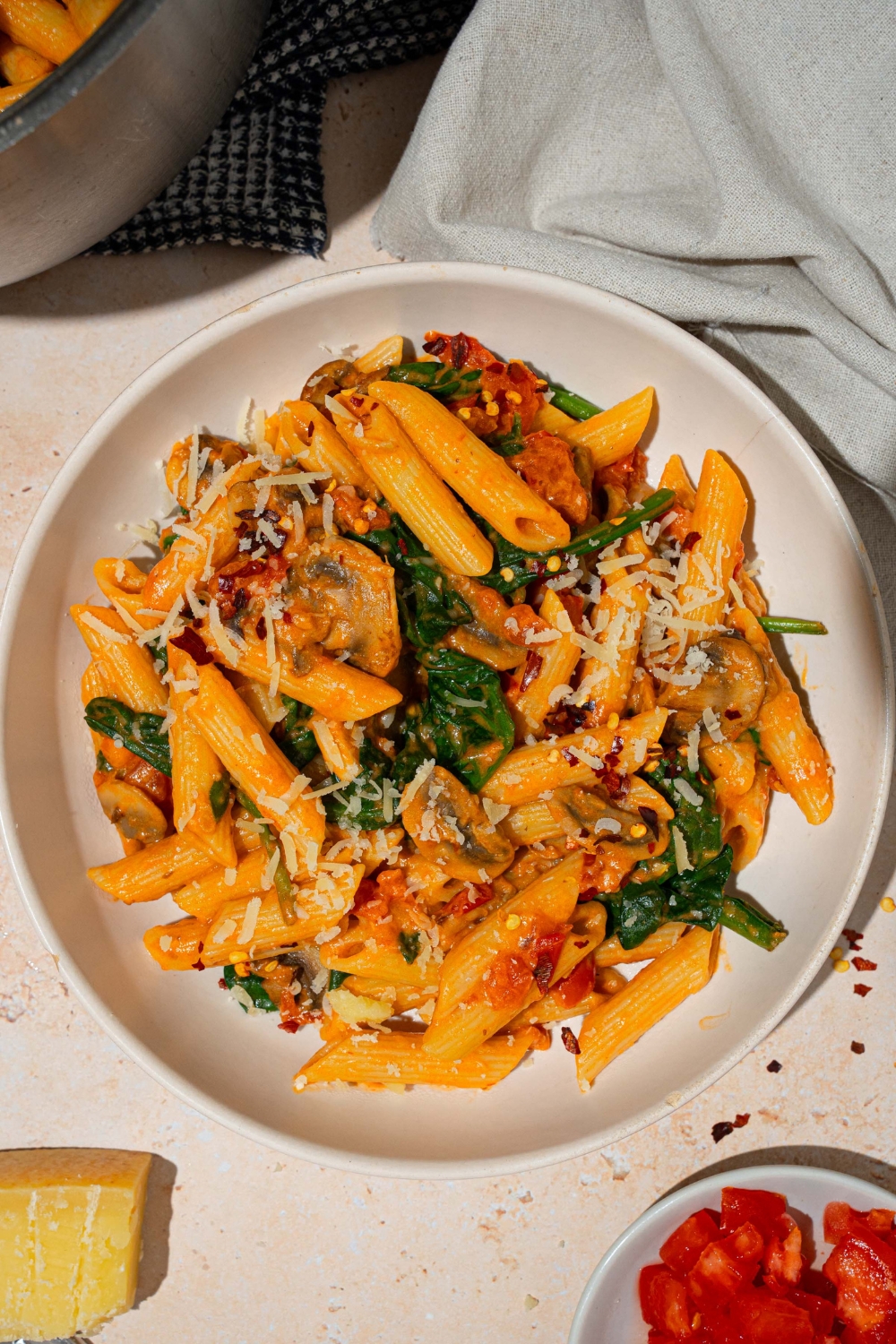 A white plate with penne rosa tossed in spinach and mushrooms and garnished with shredded parmesan. The plate is on a tan counter with a white cloth napkin and pot of penne rosa.