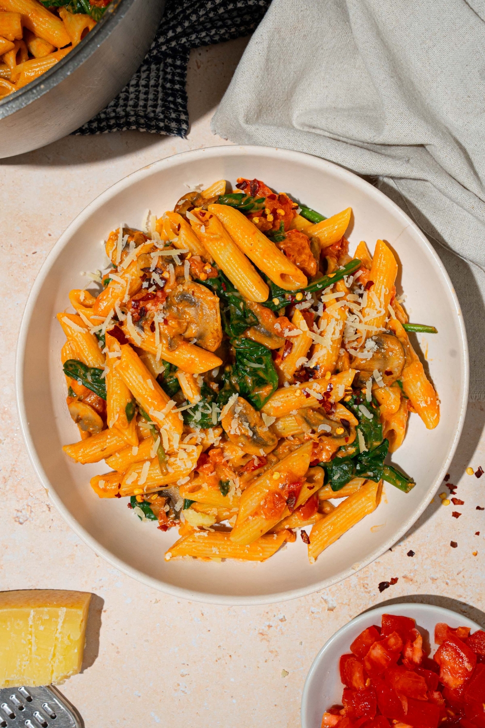 A white plate with penne rosa tossed in spinach and mushrooms and garnished with shredded parmesan. The plate is on a tan counter with a white cloth napkin and pot of penne rosa.