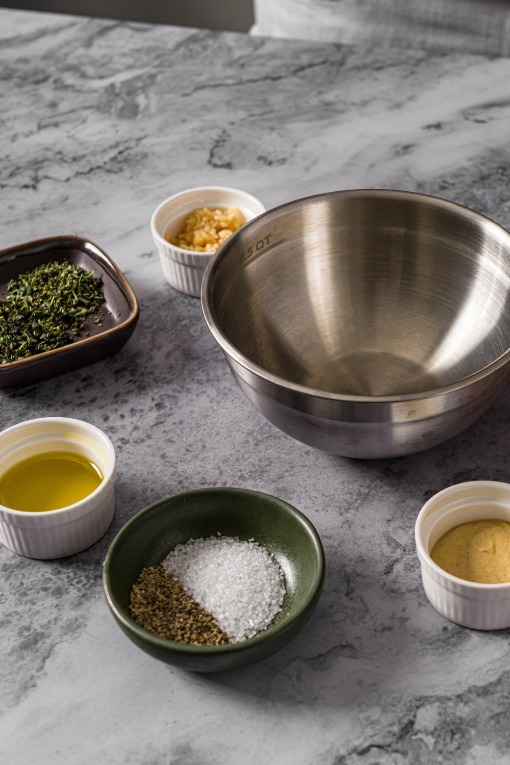 Several bowls in various sizes containing ingredients to make a seasoned mustard mixture for a rack of pork including mustard, garlic, olive oil, and seasonings.