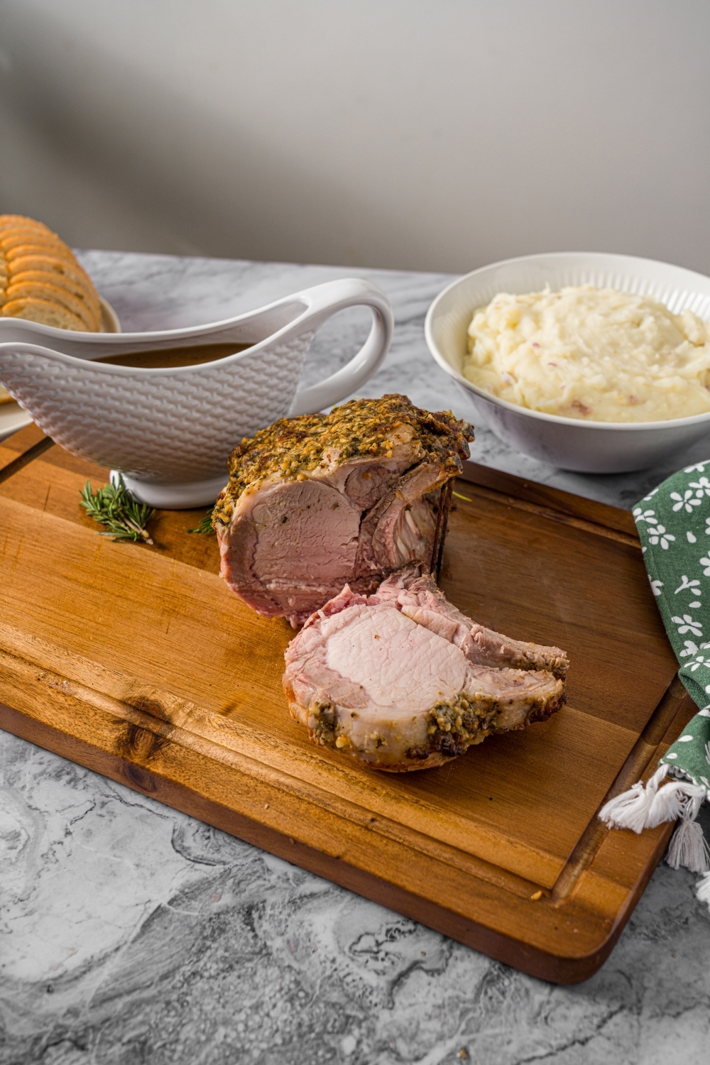 A wooden cutting board with a whole rack of pork topped with a seasoned mustard mixture. The rack has been sliced. The board is on a marble counter with a bowl of mashed potatoes and gravy boat of gravy.