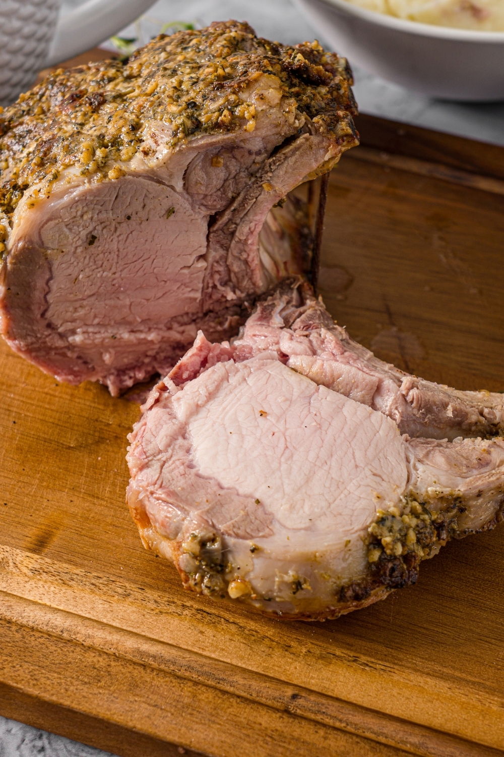 A wooden cutting board with a whole rack of pork topped with a seasoned mustard mixture. The rack has been sliced.