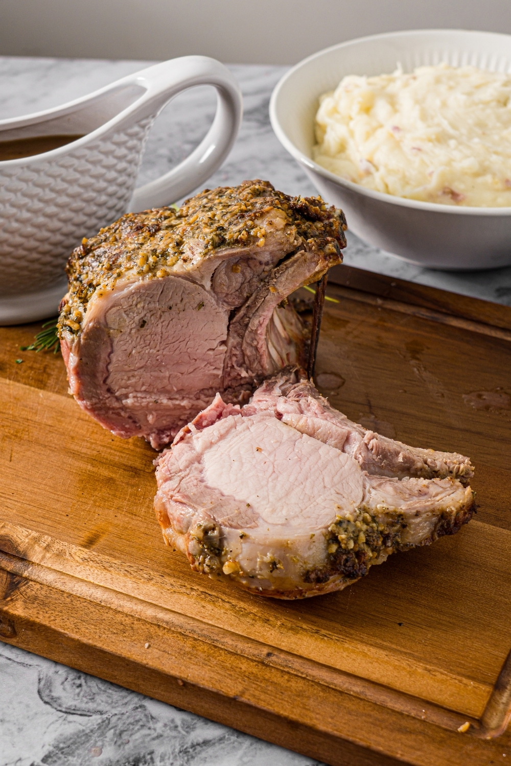 A wooden cutting board with a whole rack of pork topped with a seasoned mustard mixture. The rack has been sliced. The board is on a marble counter with a bowl of mashed potatoes and gravy boat with gravy.