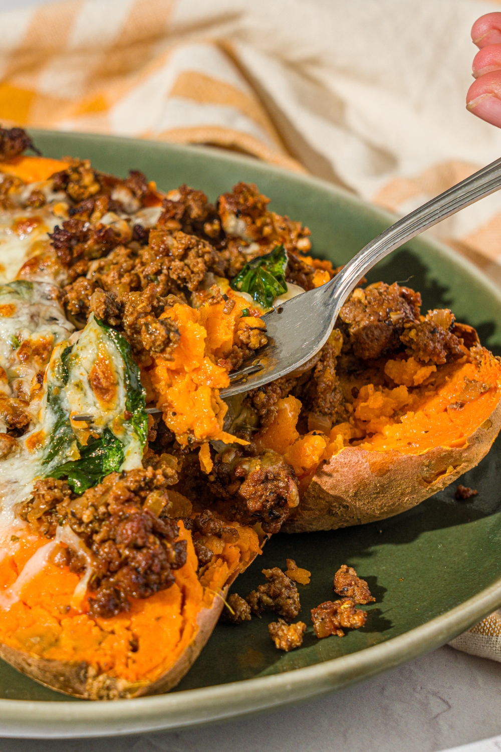 A green plate with a loaded sweet potato sliced open and stuffed with a beef mixture and topped with melted cheese. There is a fork taking a bite from the potato. The plate is on a white counter with a yellow checkered napkin.