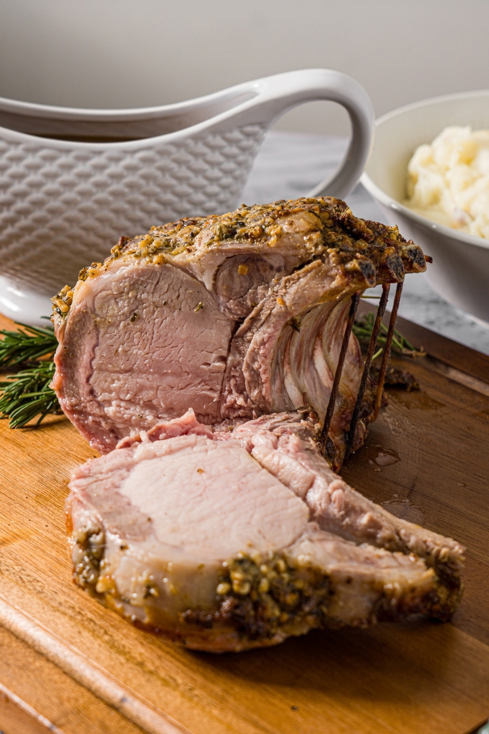 A wooden cutting board with a whole rack of pork topped with a seasoned mustard mixture. The rack has been sliced. The board is on a marble counter with a gravy boat of gravy and a bowl of mashed potatoes.