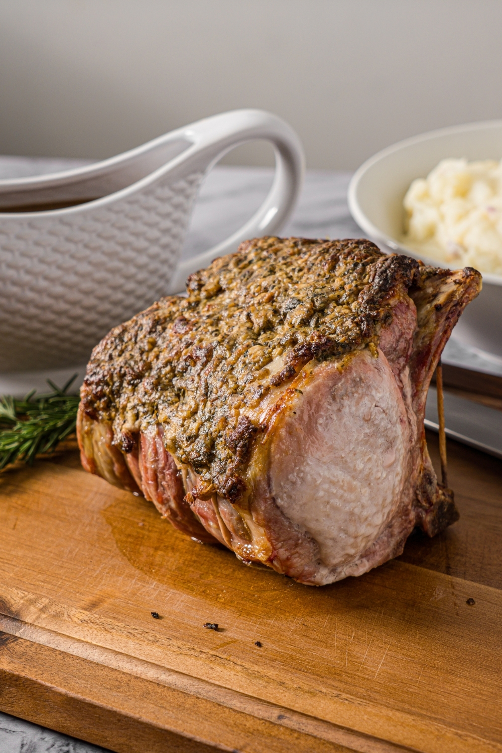 A whole rack of pork with a seasoned mustard mixture. The pork is on a wooden board on a marble counter with a bowl of mashed potatoes and gravy boat of gravy.