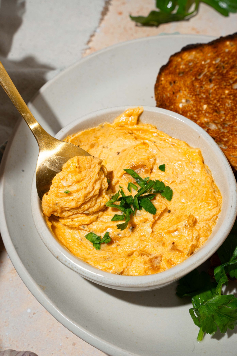 A white bowl with garlic dip garnished with fresh parsley with a spoon in the dip. The bowl is on a white plate with toasted bread. The plate is on a tan counter with a white cloth napkin.
