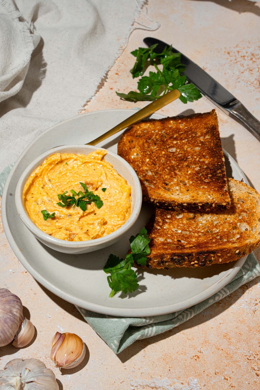 A white bowl with garlic dip garnished with fresh parsley. The bowl is on a white plate with a spoon and toasted bread. The plate is on a tan counter with a white cloth napkin.