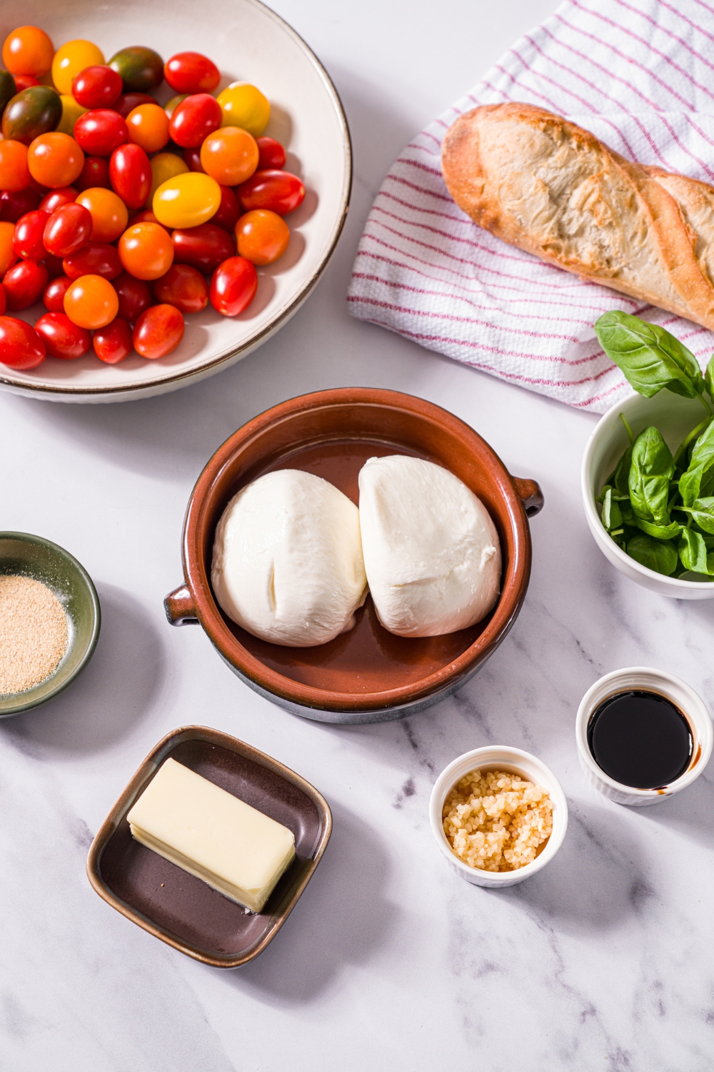 A marble counter with small bowls containing ingredients to make roasted tomato burrata dip including burrata, cherub tomatoes, fresh basil, butter, garlic, balsamic vinegar, and baguette.