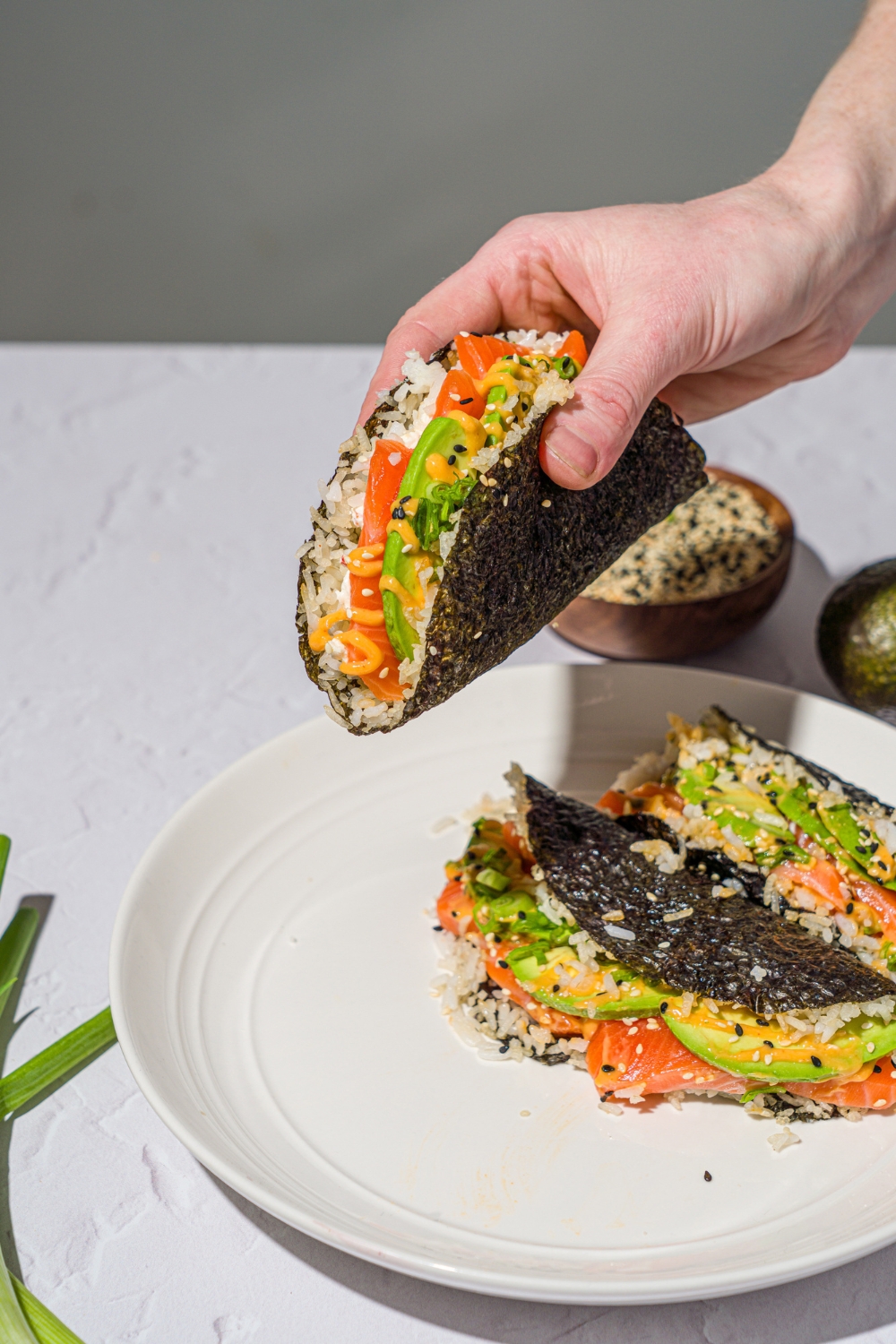 A hand holding a salmon sushi taco made with sushi rice, avocado, sriracha mayo, and cream cheese wrapped in a nori sheet and garnished with sesame seeds. There is a plate of sushi tacos on a white counter with a bowl of sesame seeds.