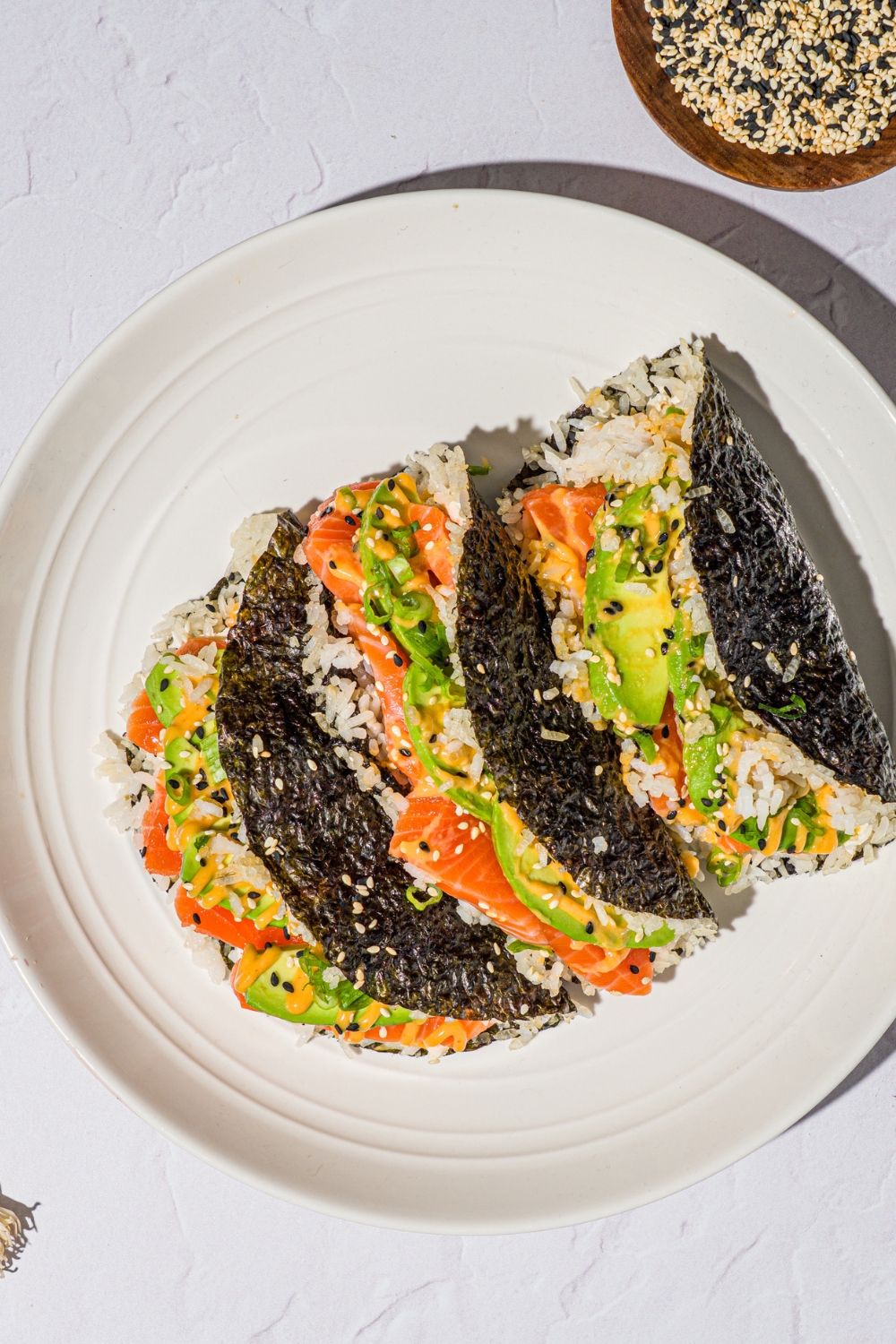 A white plate with salmon sushi tacos made with sushi rice, cream cheese, avocado, and sriracha mayo wrapped in a nori sheet and garnished with sesame seeds. The plate is on a white counter with a small bowl of sesame seeds.