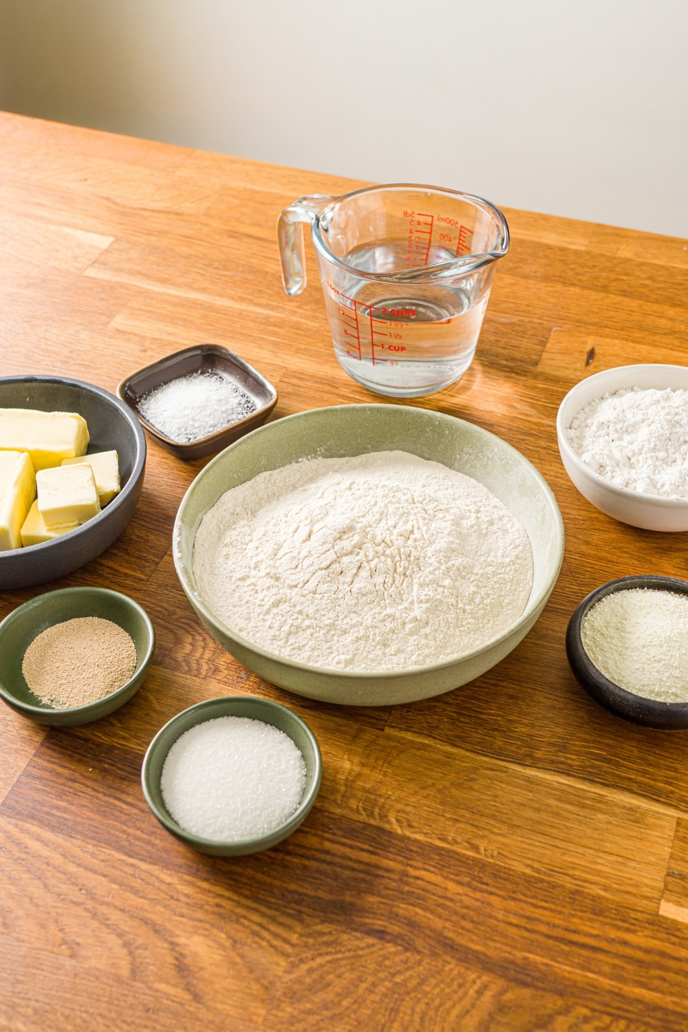 A wooden counter with several bowls in various sizes containing ingredients to make salt bread including bread dough, cake flour, water, butter, salt, milk powder, dry yeast, and more.