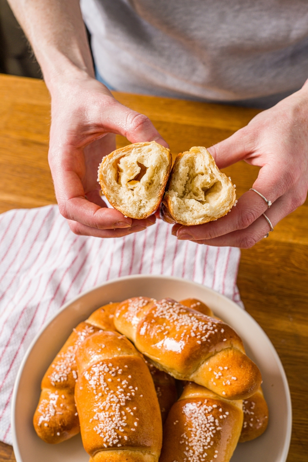 Hands holding a piece of salt bread split in half. There is a bowl of salt bread on a wooden counter with a red striped napkin.