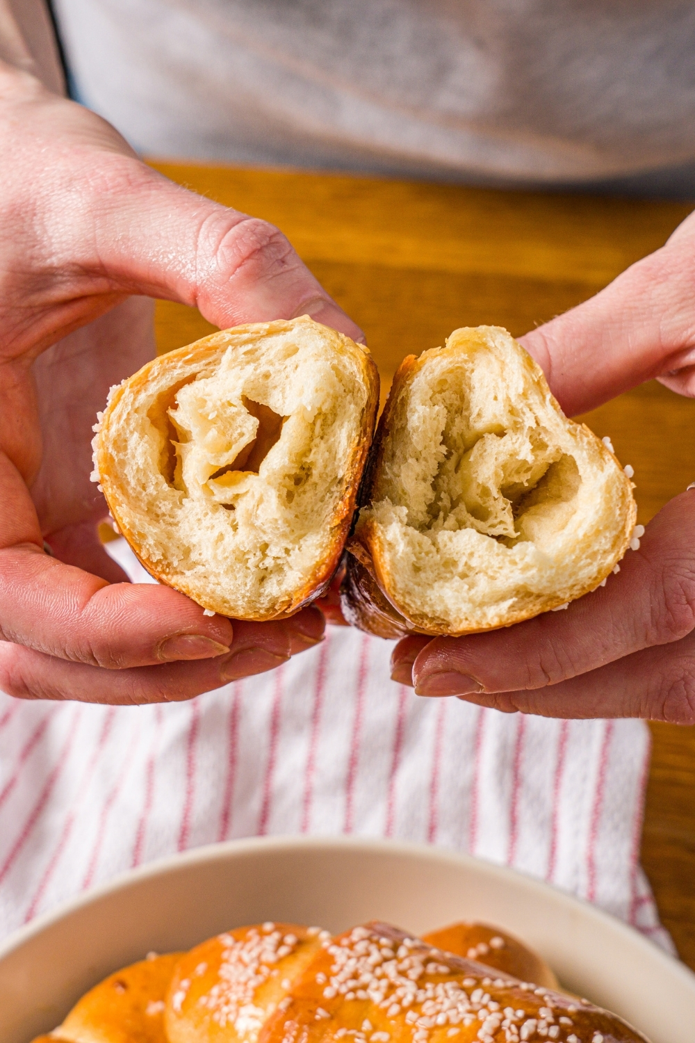 Hands holding a piece of salt bread split in half. There is a bowl of salt bread on a wooden counter with a red striped napkin.
