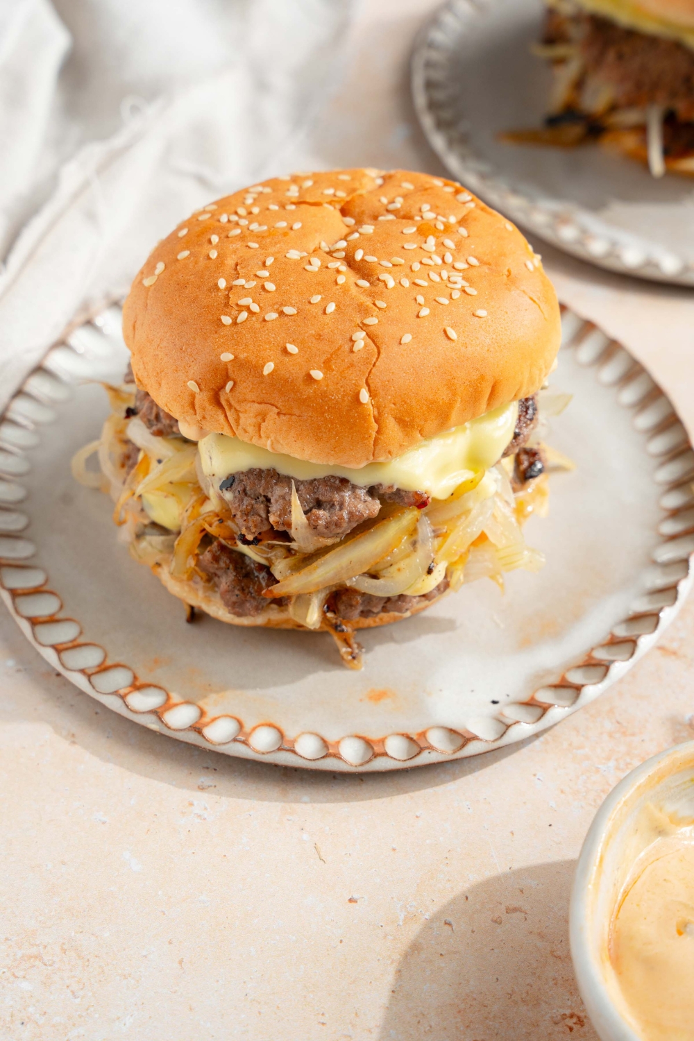 A white plate with a smash burger with onions. The plate is on a tan counter with a white cloth napkin and an additional plate with a burger.