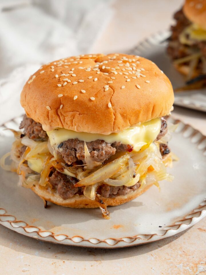 A white plate with a smash burger with onions. The plate is on a tan counter with a white cloth napkin and an additional plate with a burger.