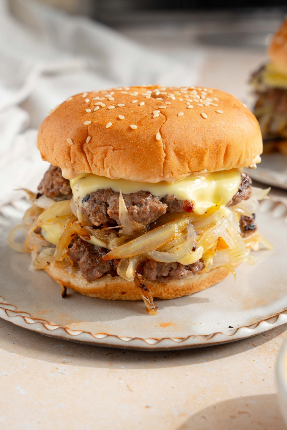 A white plate with a smash burger with onions. The plate is on a tan counter with a white cloth napkin and an additional plate with a burger.