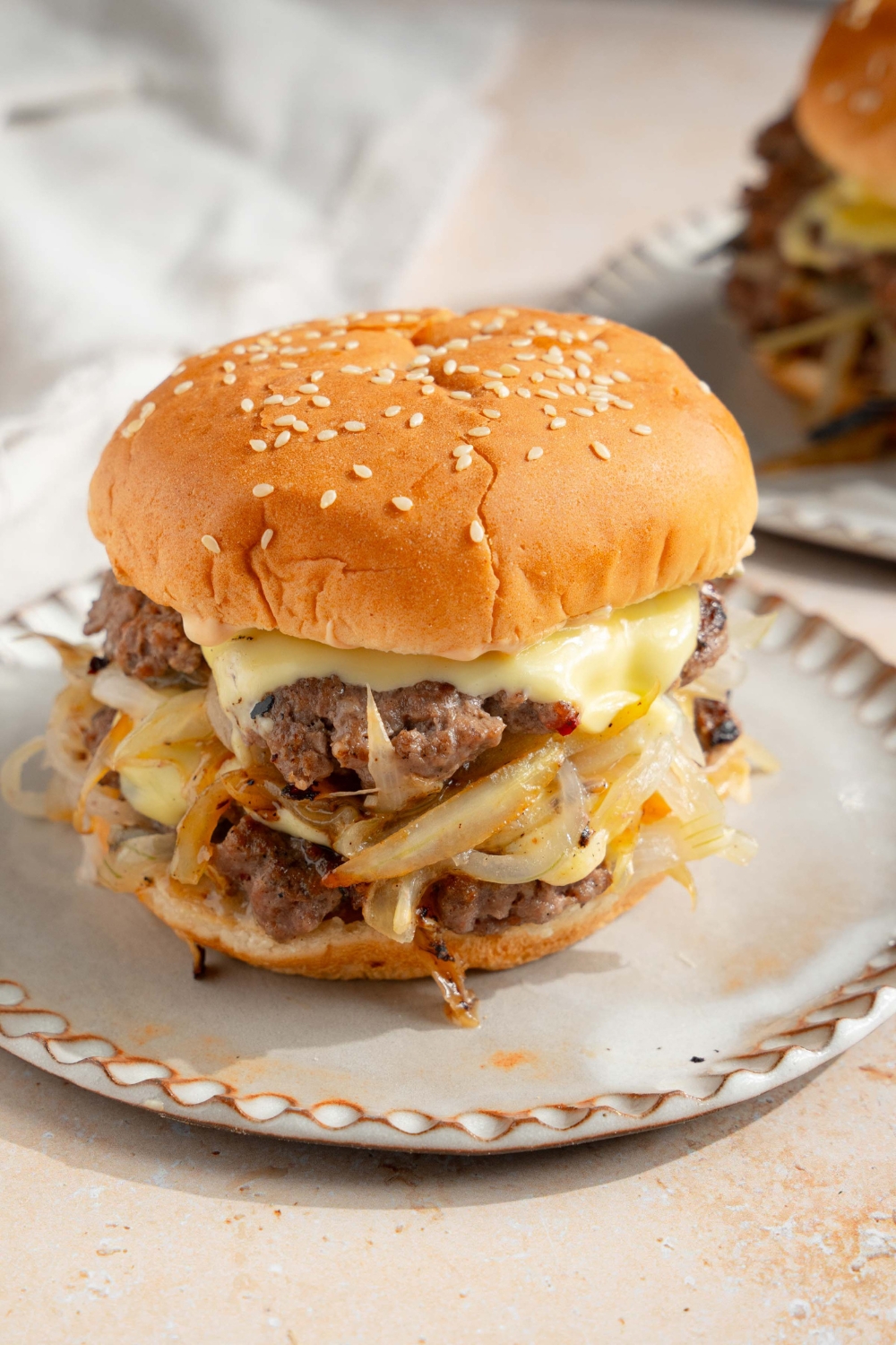 A white plate with a smash burger with onions. The plate is on a tan counter with a white cloth napkin and an additional plate with a burger.