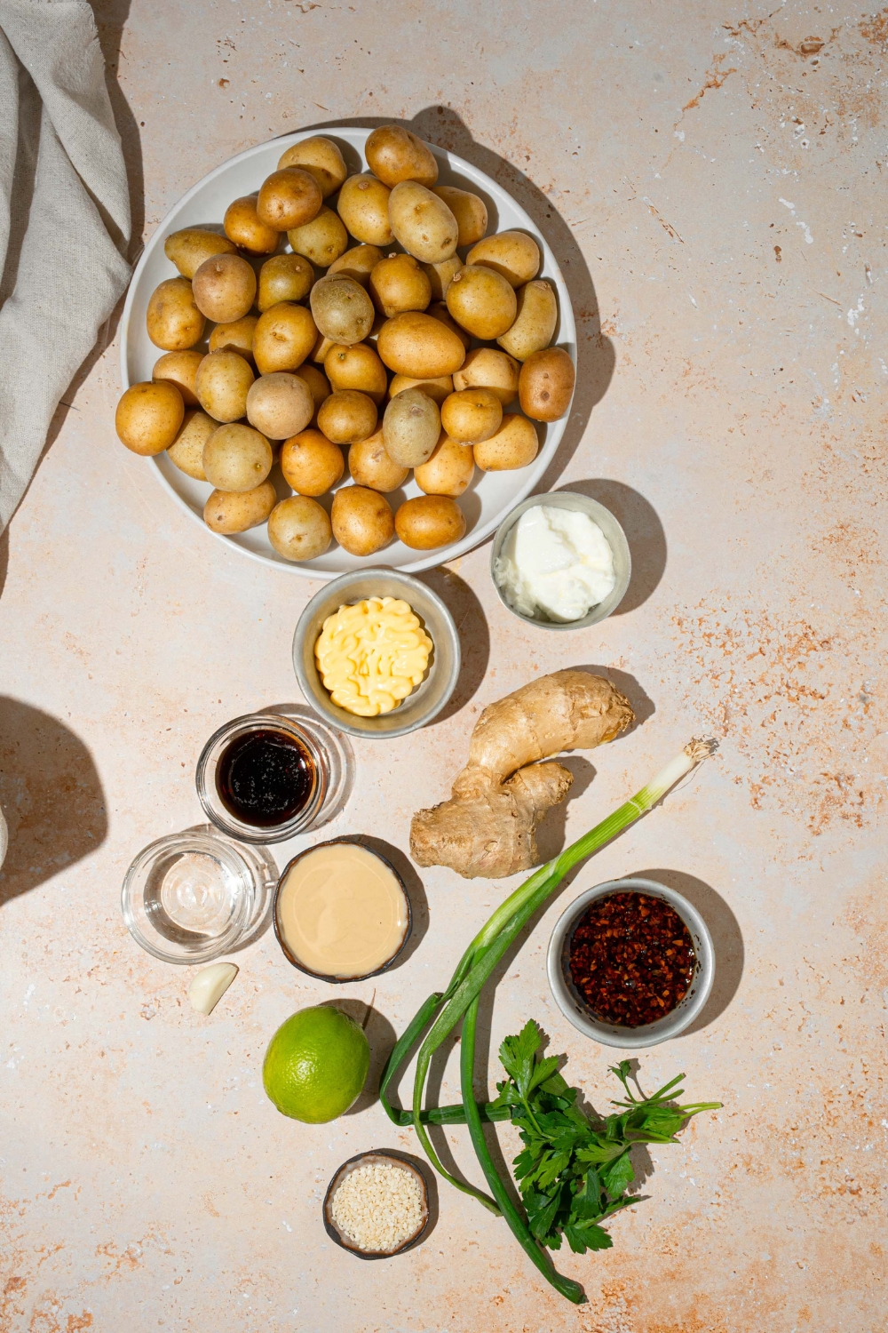 An overhead shot of several bowls in various sizes containing ingredients to make smashed potato salad including baby potatoes, greek yogurt, tahini, garlic, chili crisp, soy sauce, ginger, green onion, and sesame seeds.