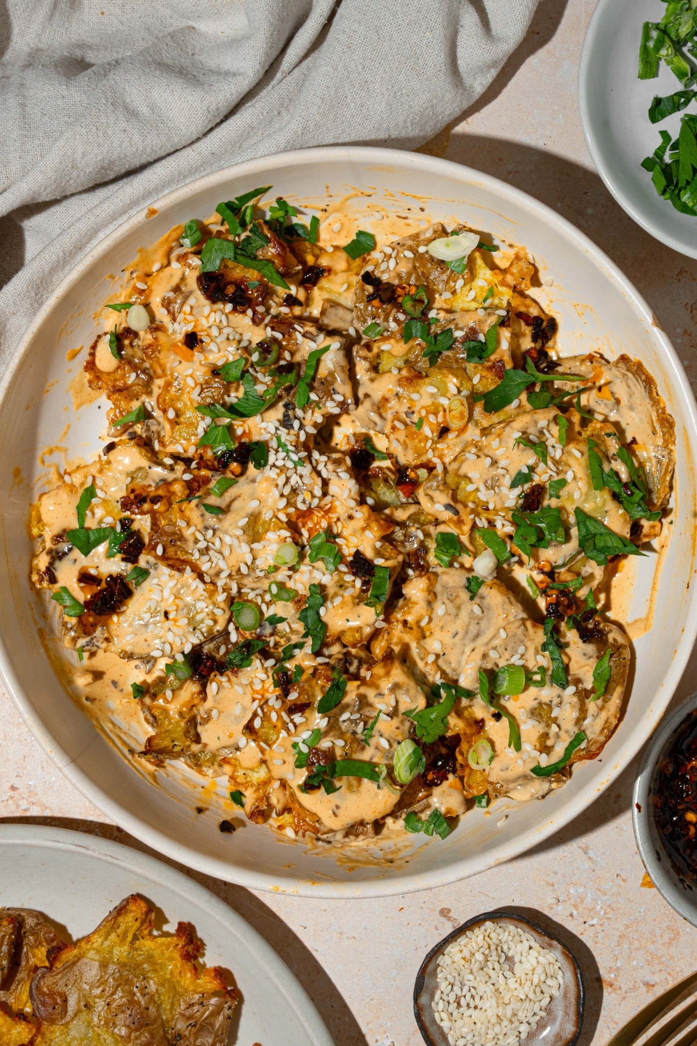 A white bowl with smashed potato salad tossed in chili crisp tahini dressing garnished with sesame seeds and sliced green onion. The bowl is on a tan counter with an additional plate of crispy potatoes and small bowls of garnishes.