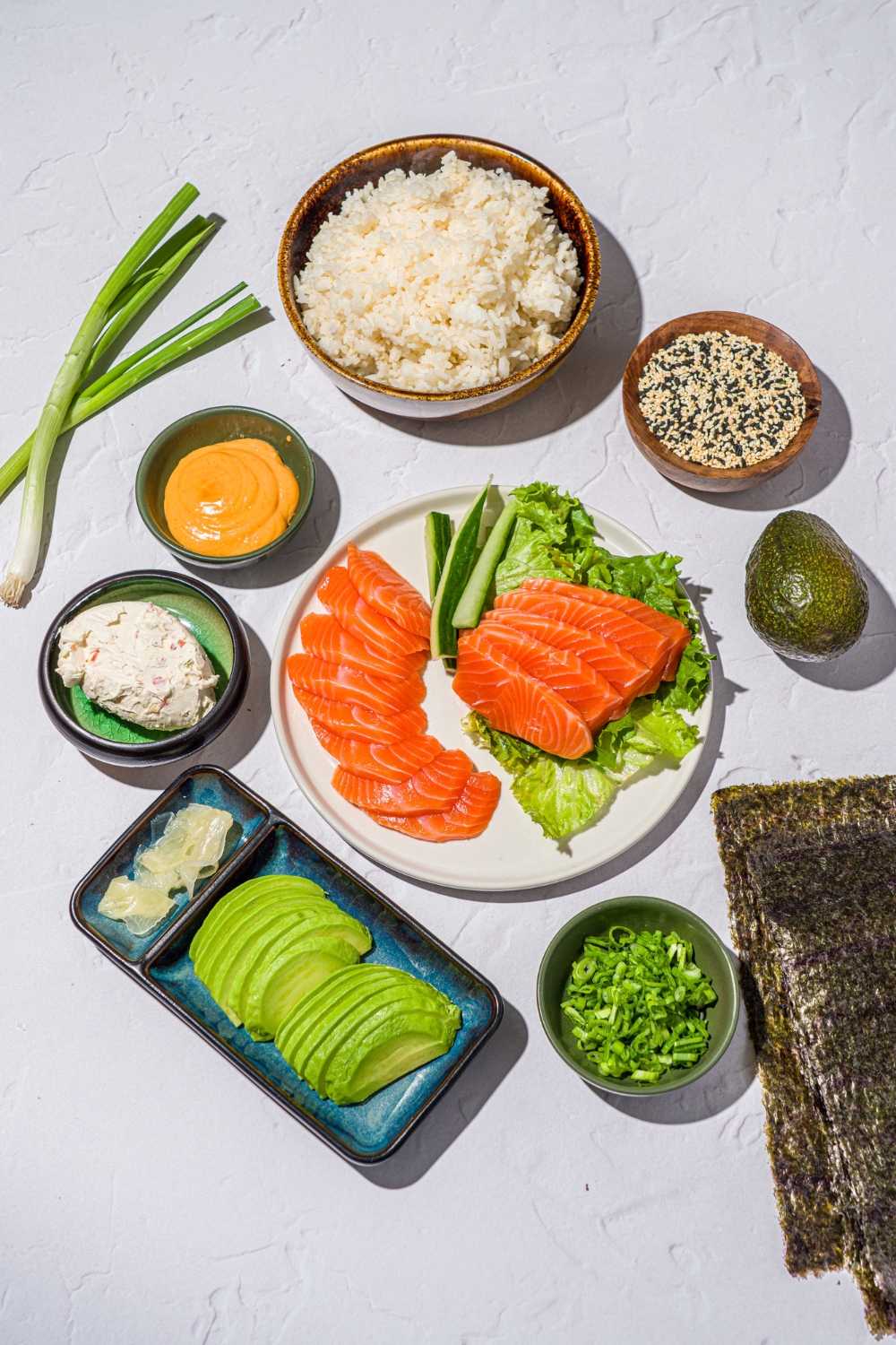 A white counter with several bowls in various sizes containing ingredients to make sushi tacos including nori sheets, sushi rice, salmon, avocado, cream cheese, green onion, sriracha mayo, and sesame seeds.