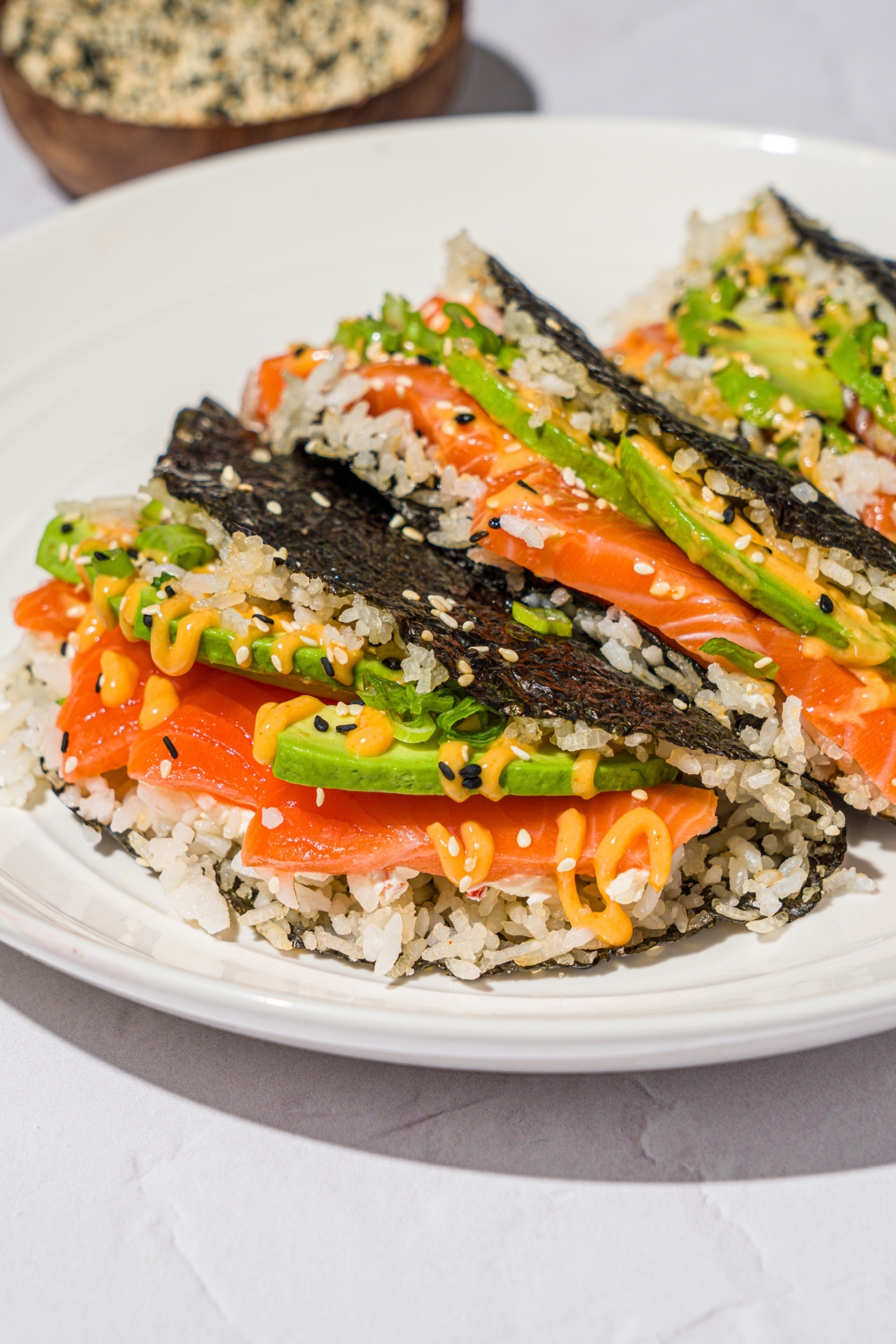 A white plate with salmon sushi tacos made with sushi rice, cream cheese, avocado, and sriracha mayo wrapped in a nori sheet and garnished with sesame seeds. The plate is on a white counter with a small bowl of sesame seeds.