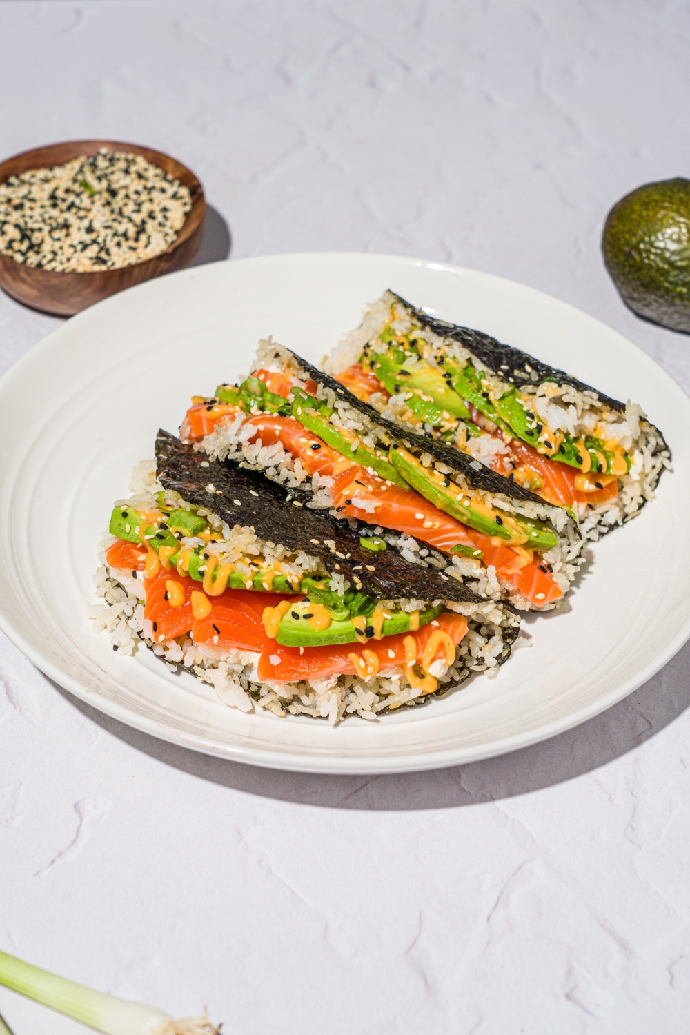 A white plate with salmon sushi tacos made with sushi rice, cream cheese, avocado, and sriracha mayo wrapped in a nori sheet and garnished with sesame seeds. The plate is on a white counter with a small bowl of sesame seeds.