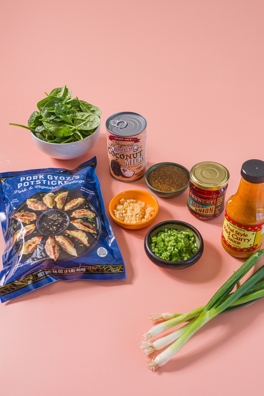 A pink counter with ingredients to make Trader Joe's chili crisp dumpling bake including a bag of Trader Joe's chili crisp dumplings, coconut milk, thai red curry sauce, soyaki, garlic, spinach, green onions, and a jar of Trader Joe's chili onion crunch.