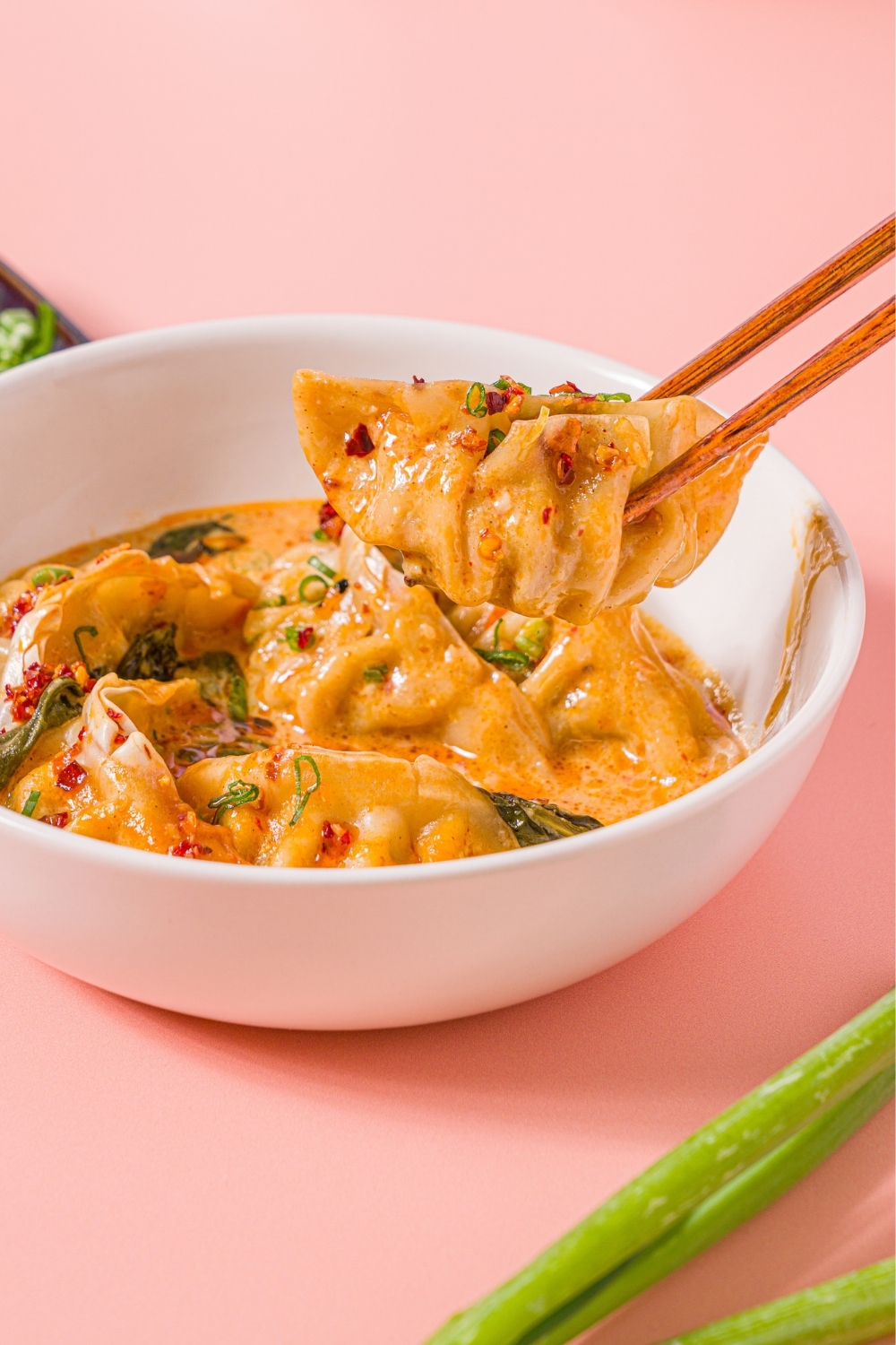 A white bowl with Trader Joe's chili crisp dumpling bake garnished with sliced green onion and Trader Joe's chili onion crunch. A pair of chopsticks is taking a dumpling from the bowl. The bowl is on a pink counter.