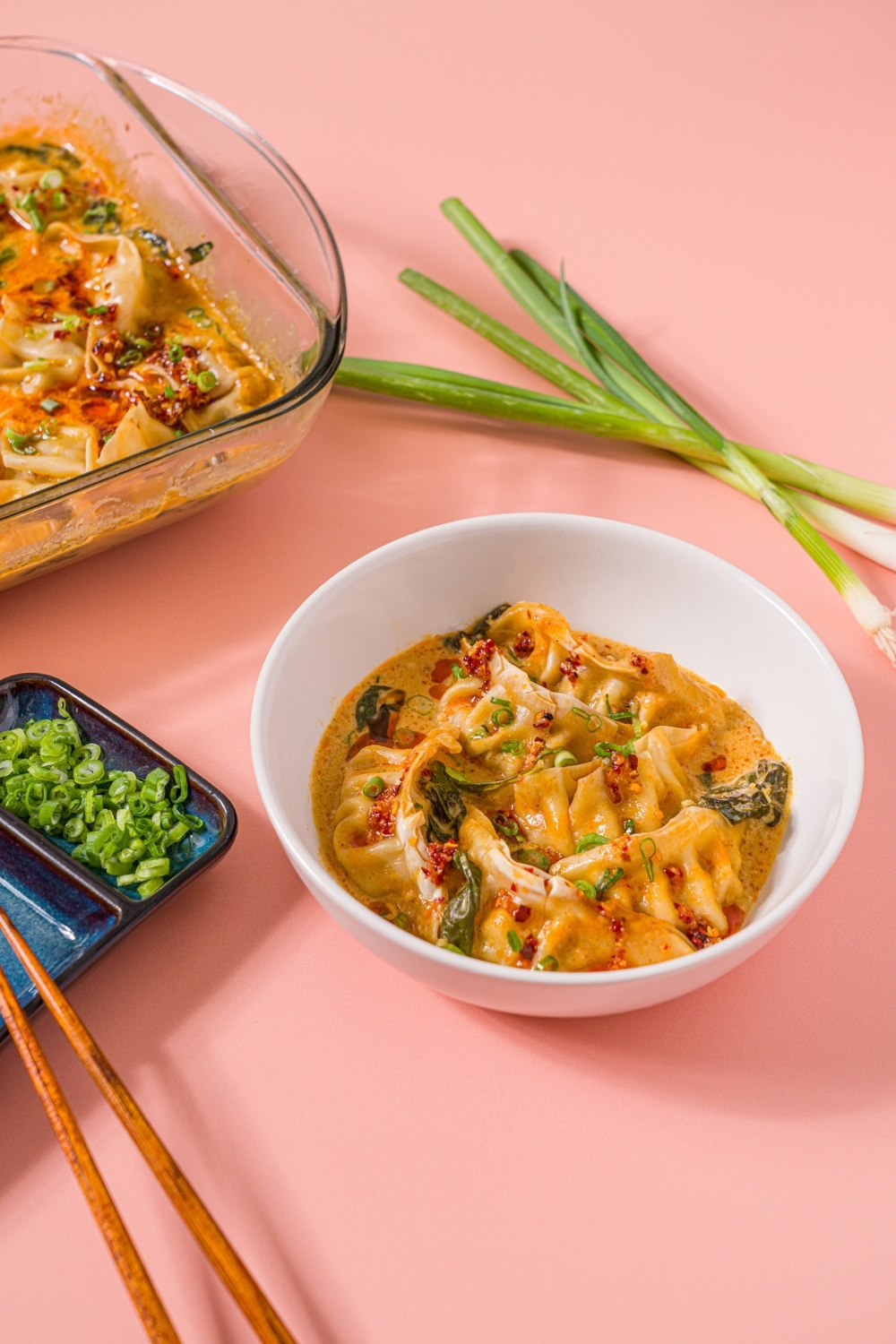 A white bowl with Trader Joe's chili crisp dumpling bake garnished with sliced green onion and Trader Joe's chili onion crunch. The bowl is on a pink counter with a pair of chopsticks and a baking dish of dumpling bake.