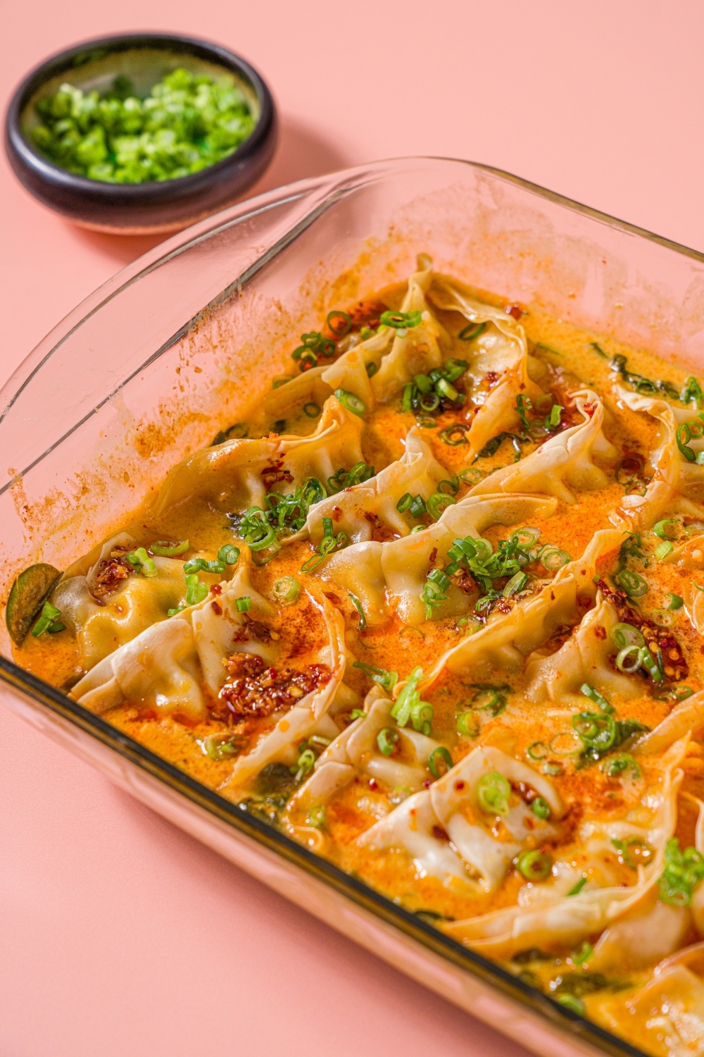 A baking dish with Trader Joe's chili crisp dumpling bake garnished with sliced green onion and Trader Joe's chili onion crunch. The dish is on a pink counter with a small bowl of sliced green onions.