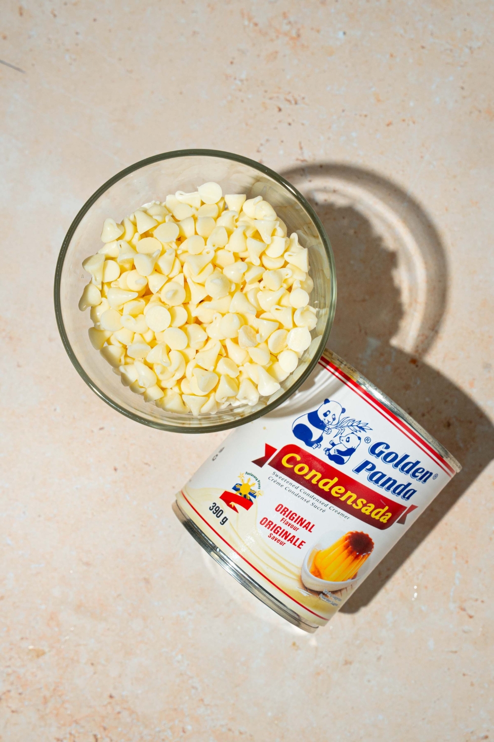 A bowl of white chocolate chips and can of condensed milk on a tan counter.