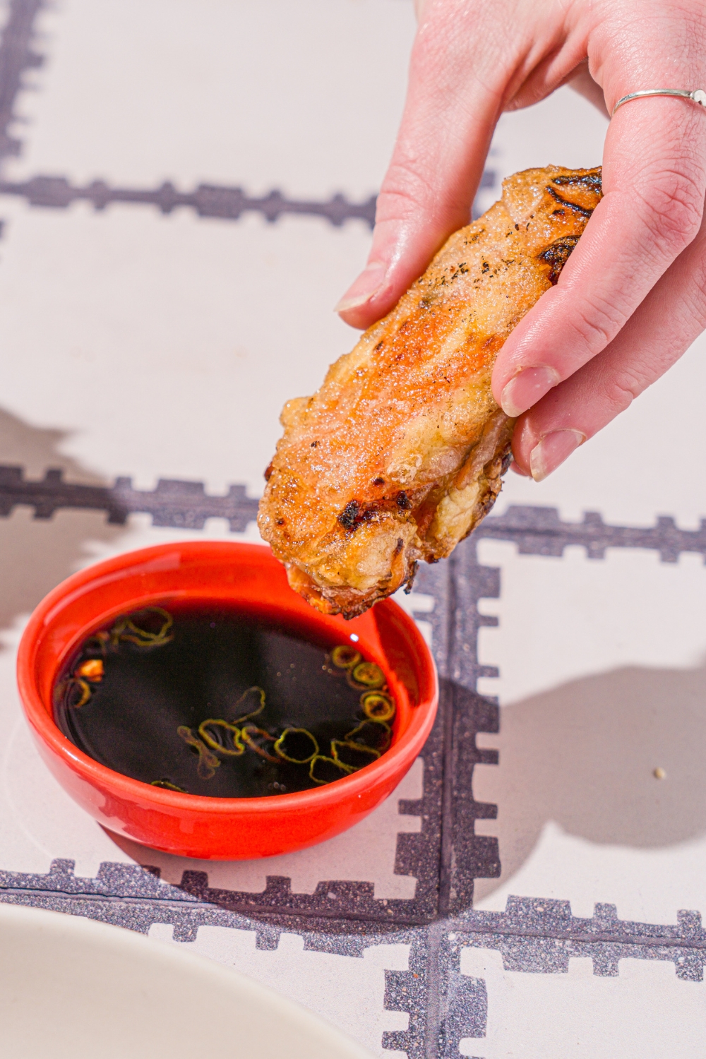 A hand dipping a veggie rice paper dumpling into a bowl of dipping sauce. The bowl is on a tiled counter.
