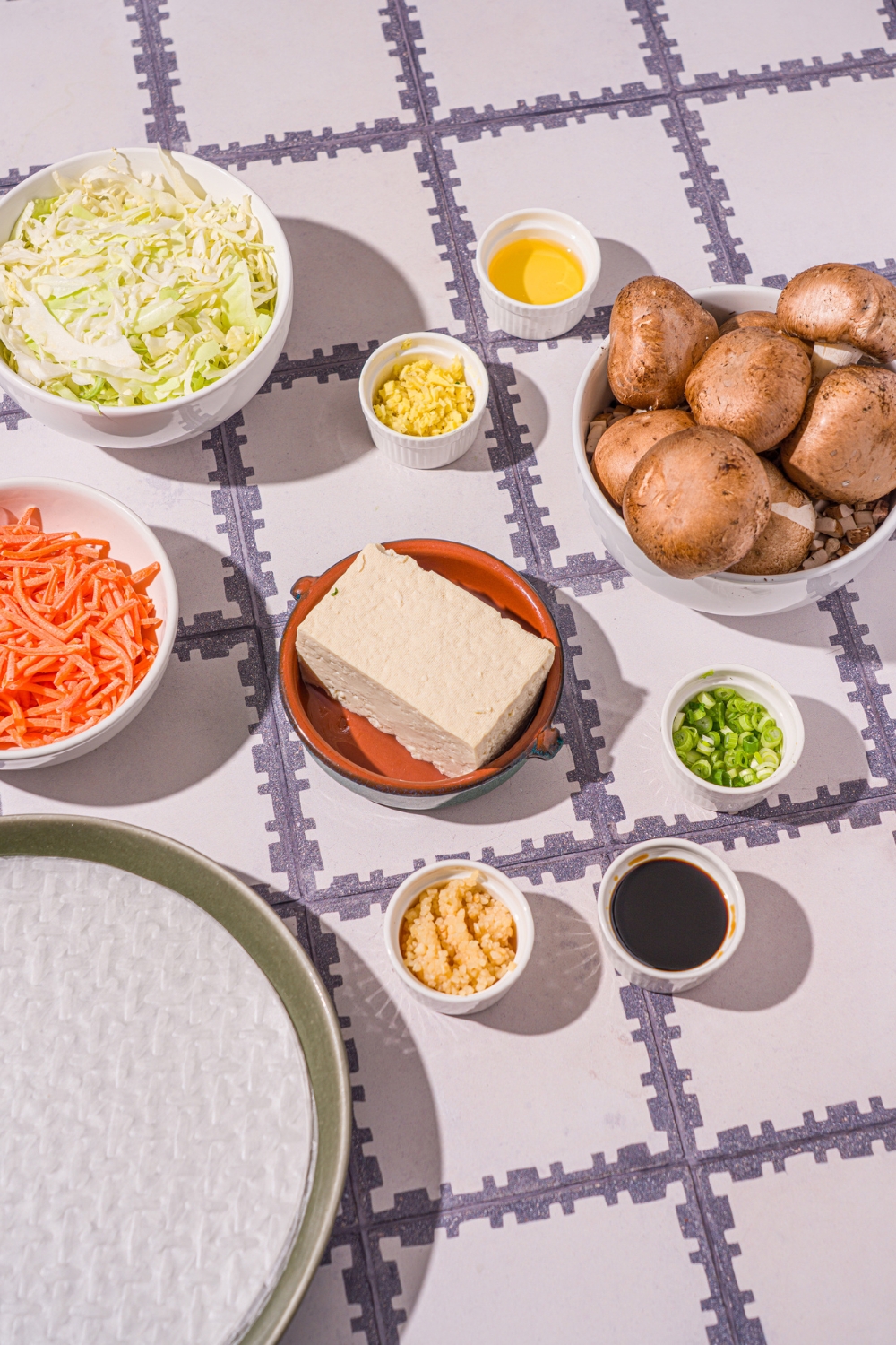 A tiled counter with several bowls in various sizes containing ingredients to make vegetable rice paper dumplings including sheets of rice paper, tofu, shredded cabbage, shredded carrots, mushrooms, garlic, sliced green onions, and more.