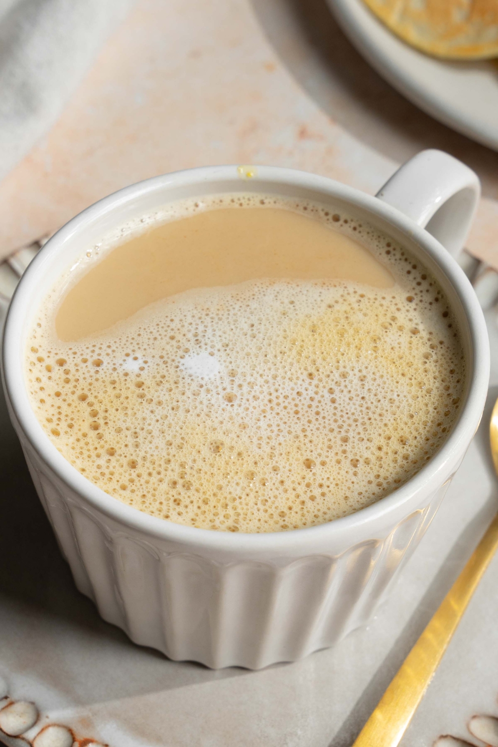 A white mug with Vietnamese egg coffee on saucer on a tan counter with a spoon.