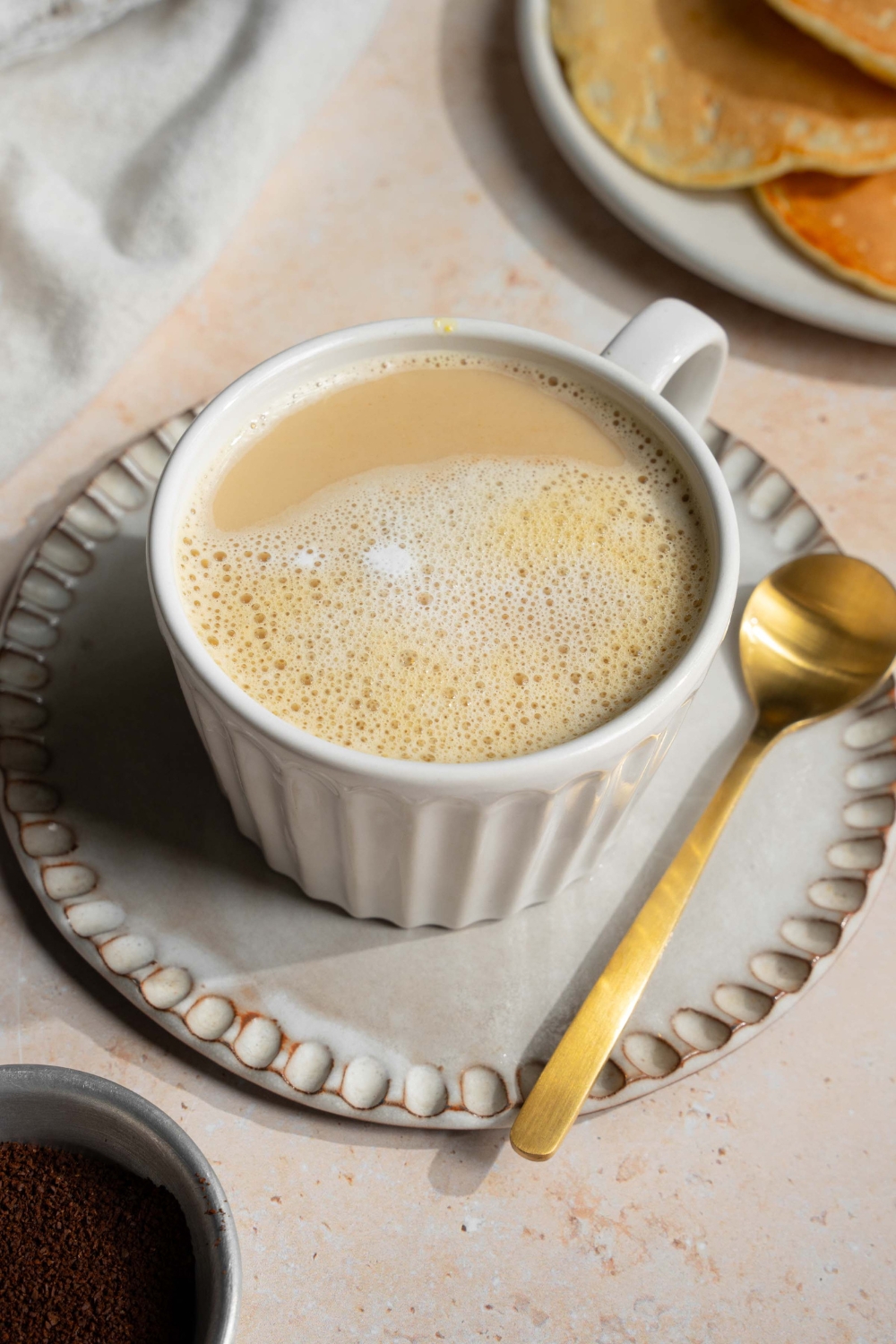 A white mug with Vietnamese egg coffee on a saucer with a spoon. The saucer is on a tan counter with a plate of pancakes and a white cloth napkin.
