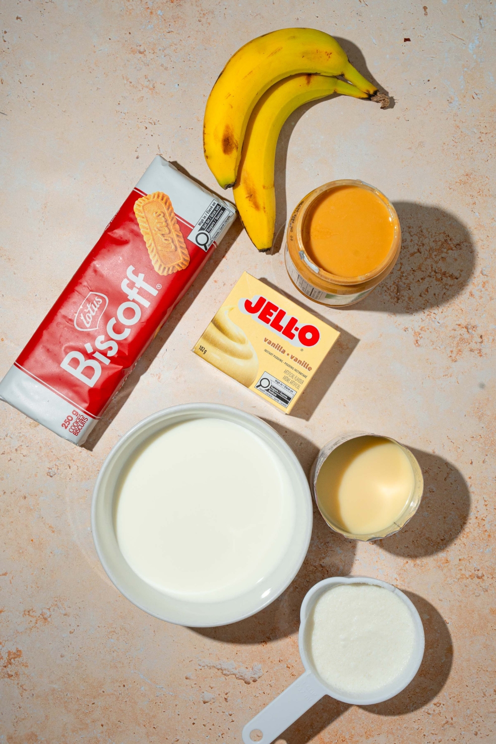 A tan counter with ingredients to make Biscoff banana pudding including bananas, Biscoff cookies, milk, cream, vanilla pudding mix, and condensed milk.